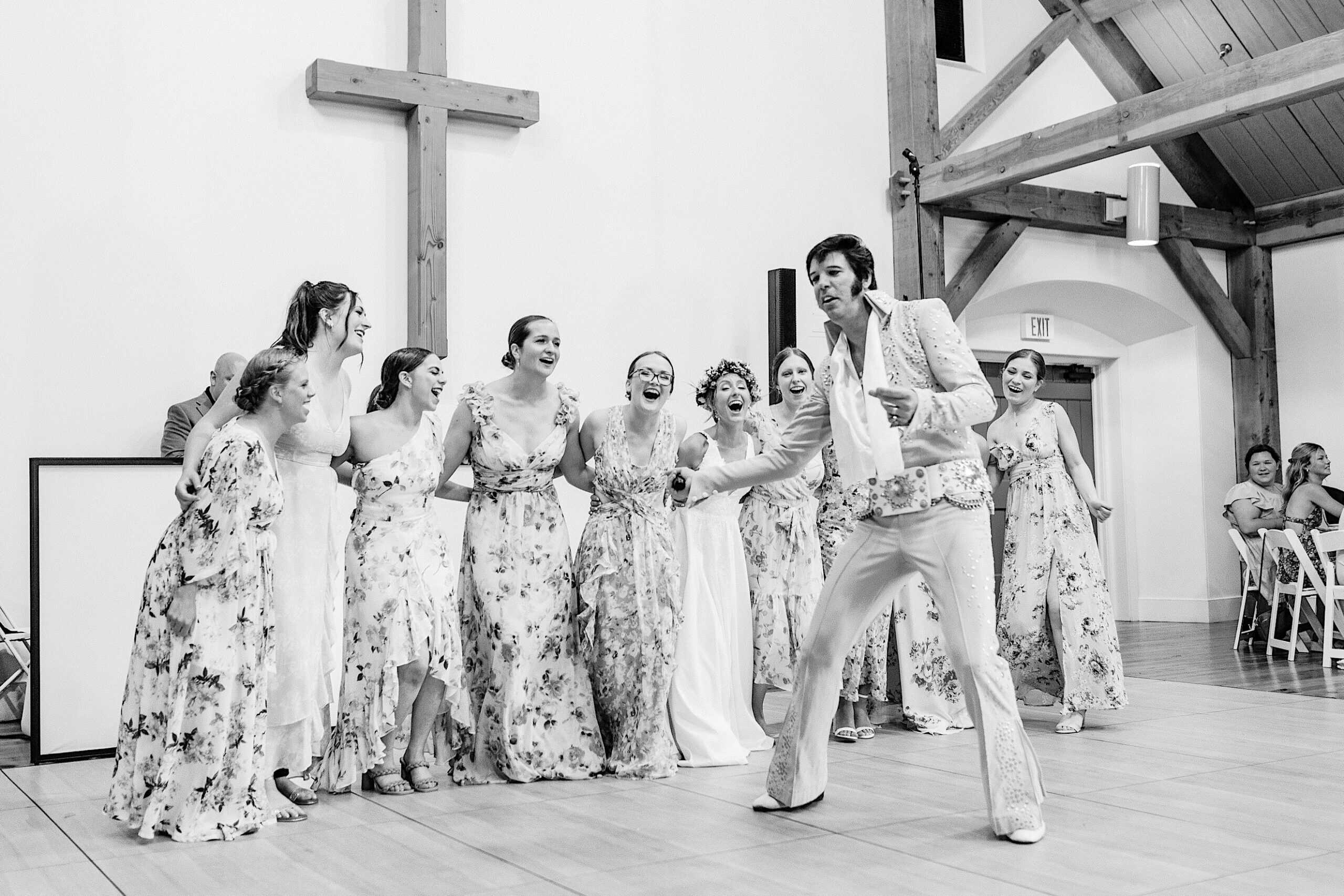 At a Summer Wedding at The High Point, a group of women in floral dresses stand together smiling and singing while a man dressed as Elvis Presley performs in front of them in a wooden-beamed hall.