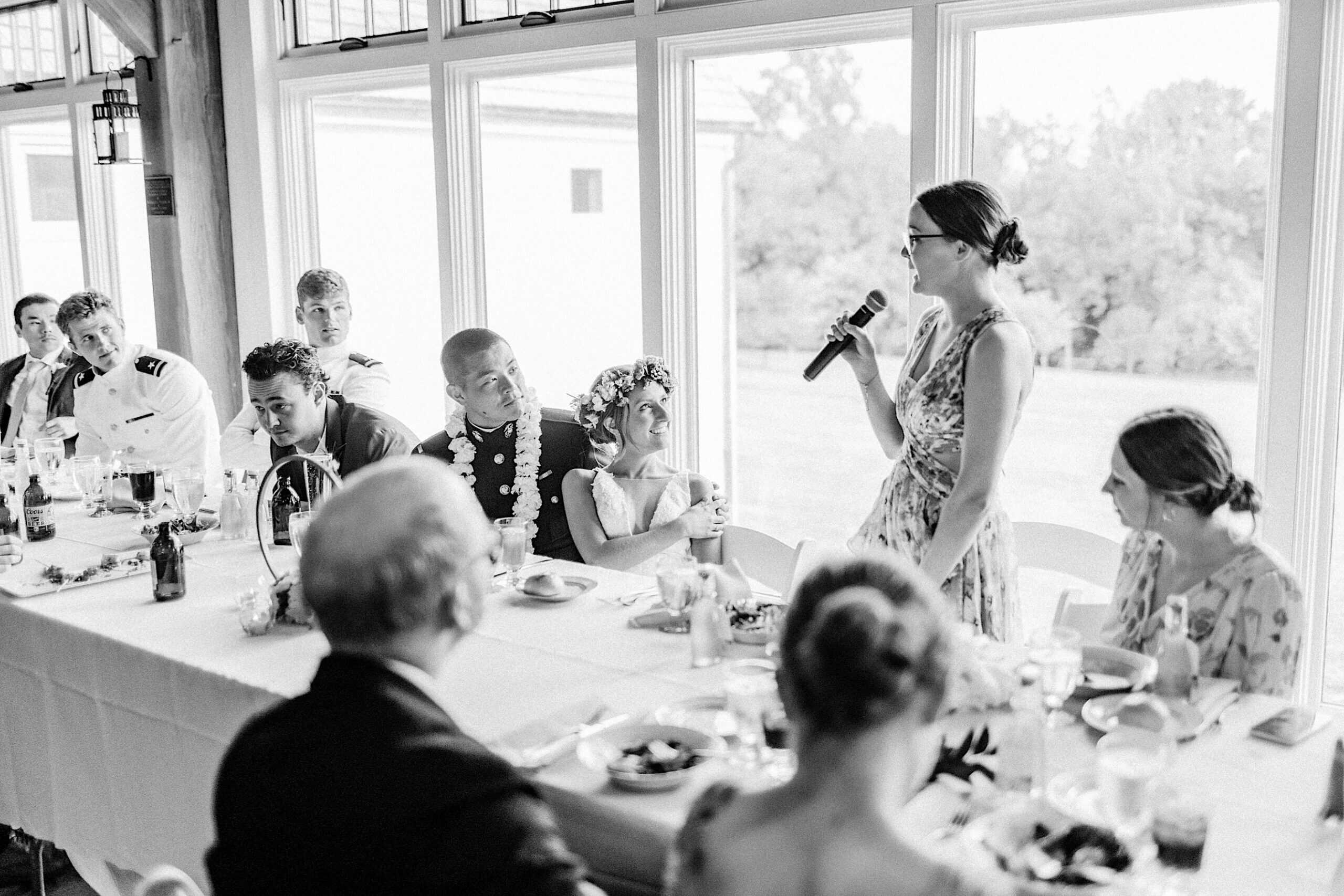At a Summer Wedding at The High Point, a woman stands and speaks into a microphone at a long table during the reception, while guests, including the bride and groom, listen attentively.