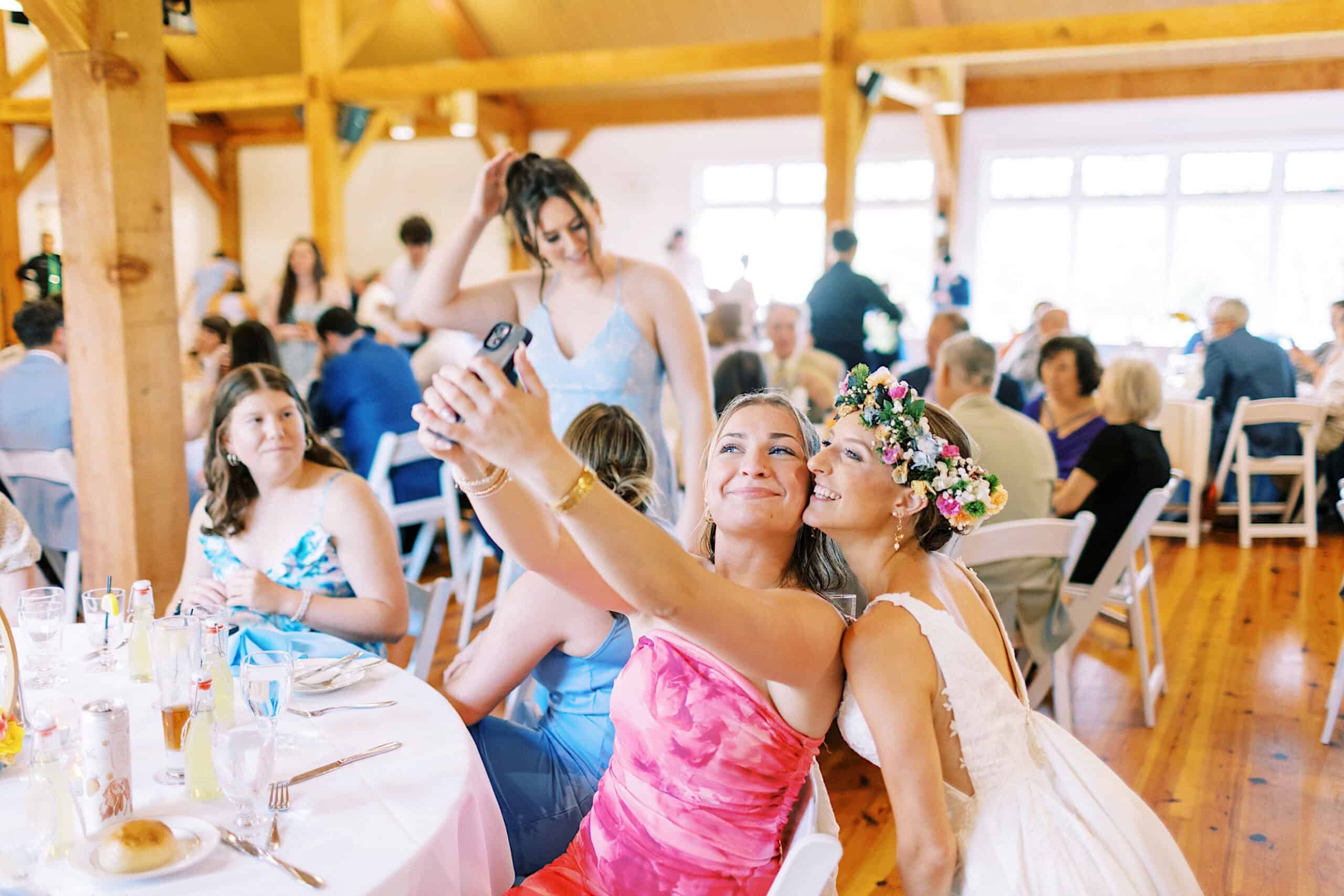 Two women seated at a table take a selfie at a lively indoor event; one in a floral crown and white dress hints at a Summer Wedding at The High Point, capturing the joyful celebration.