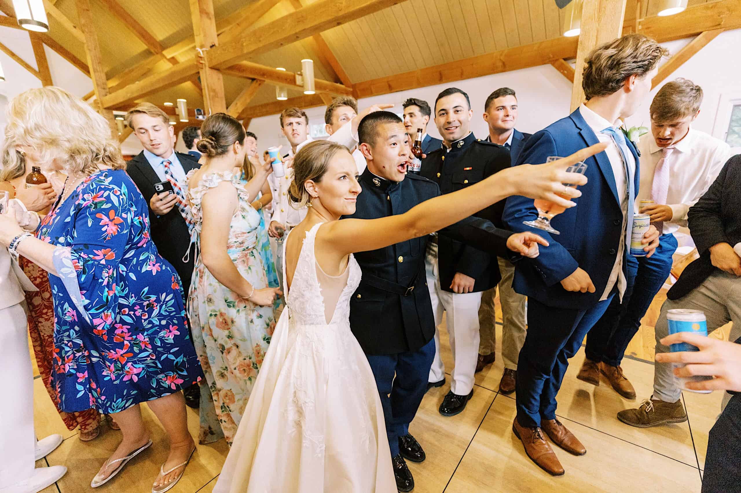 At a Summer Wedding at The High Point, a bride in a white dress takes a selfie with guests. People in formal attire, some holding drinks, gather around her in a hall with beautiful wooden beams.