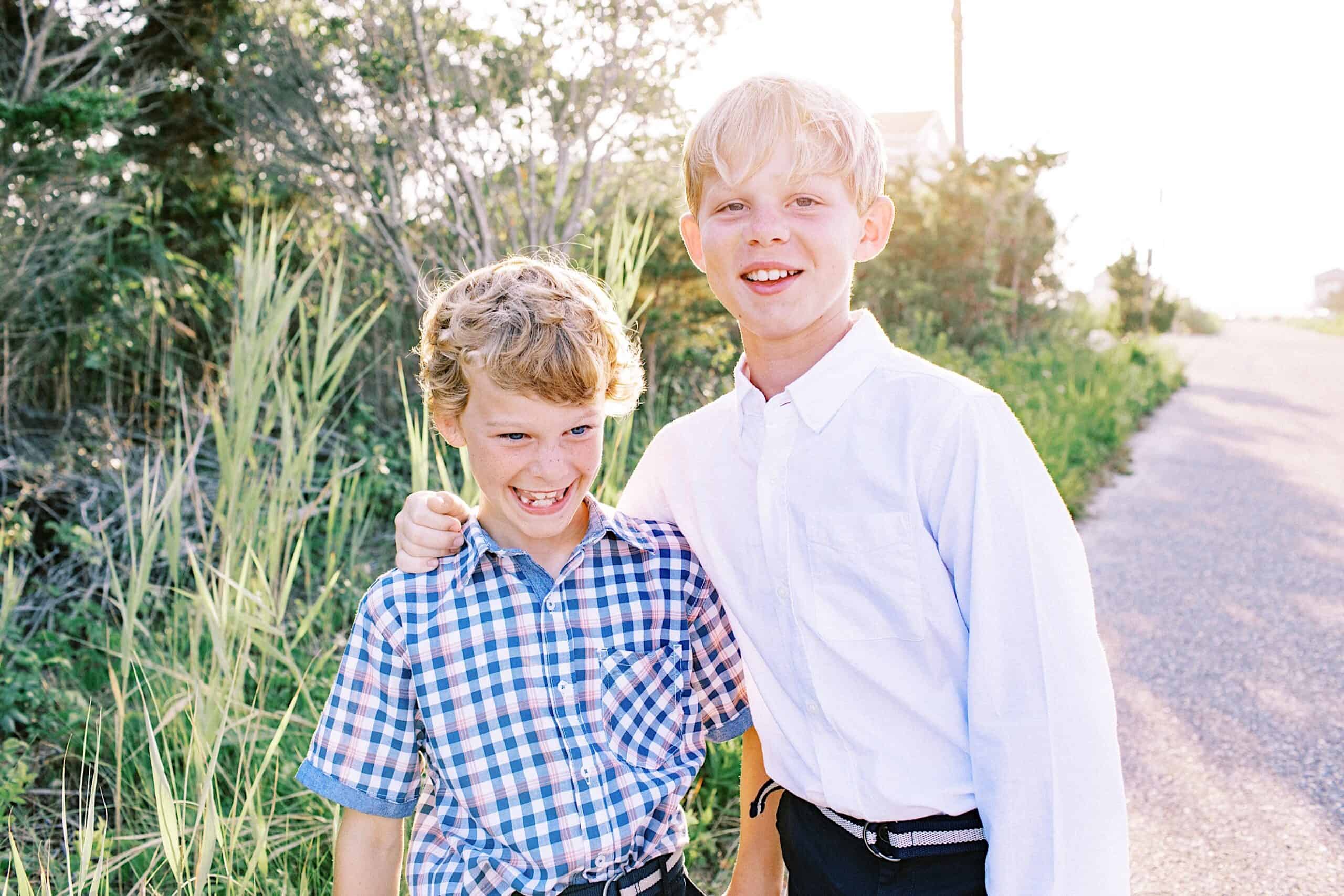 Two young boys stand outside on a sunny day, one with his arm around the other. Both are smiling; one wears a white shirt, the other a blue plaid shirt. It's a perfect scene for Jersey Shore Family Photos, with grass and a path in the background.