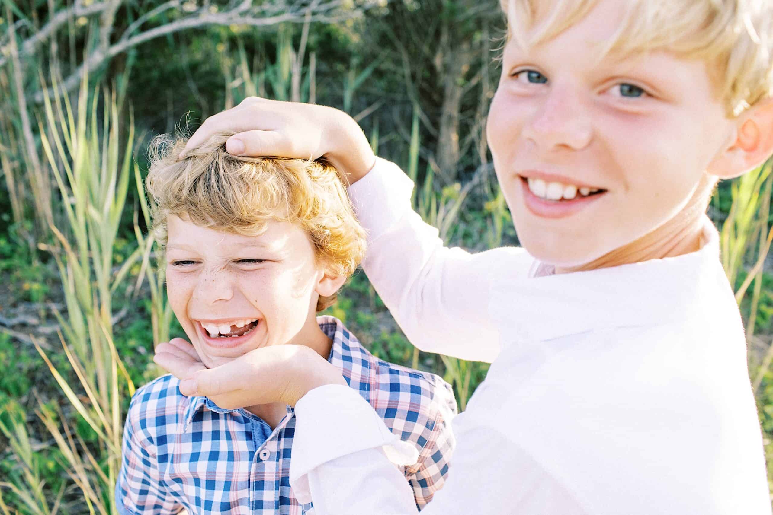 Two boys outdoors, one in a white shirt holding the other’s head and chin, both smiling and laughing. Jersey Shore Family Photos vibes shine through with tall grass and trees in the background.