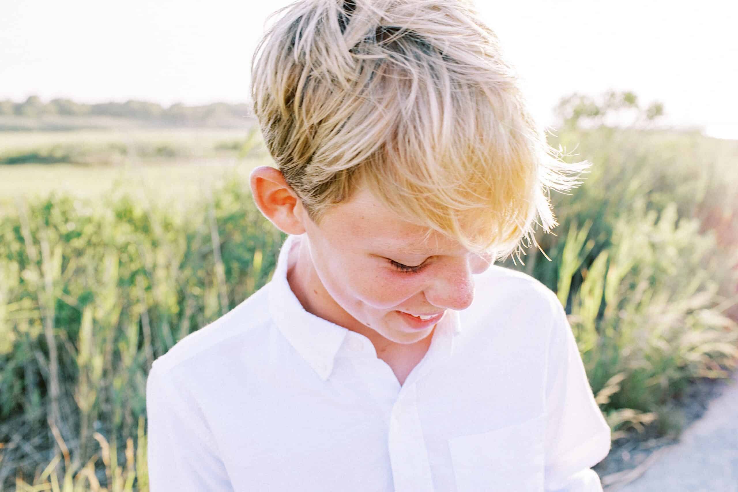 A young boy with blonde hair wearing a white shirt looks down and smiles outdoors in bright natural light, with greenery in the background—a perfect moment for Jersey Shore Family Photos.