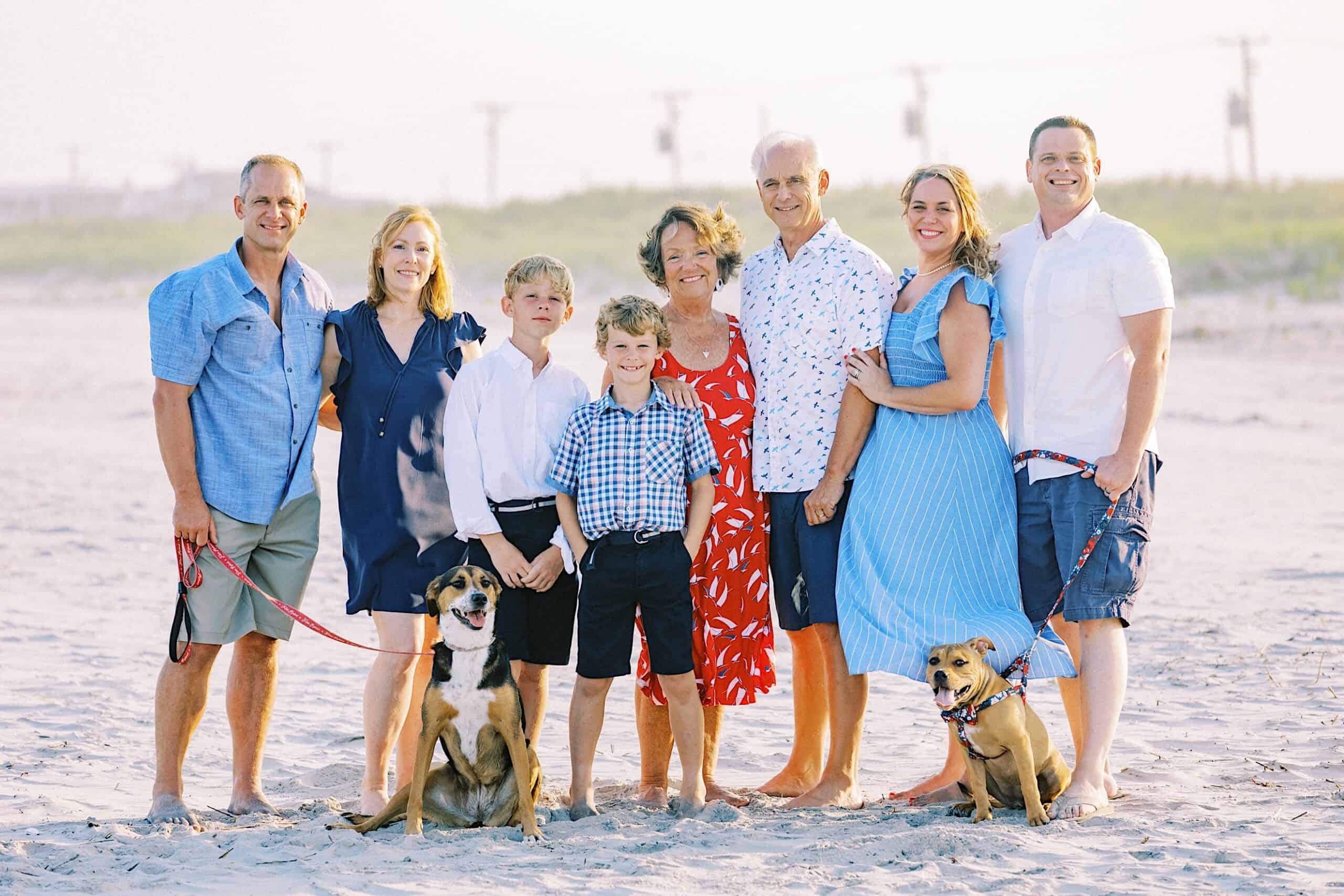 A group of eight people, including two children and two dogs, pose together on a sandy beach with grassy dunes in the background for classic Jersey Shore Family Photos.