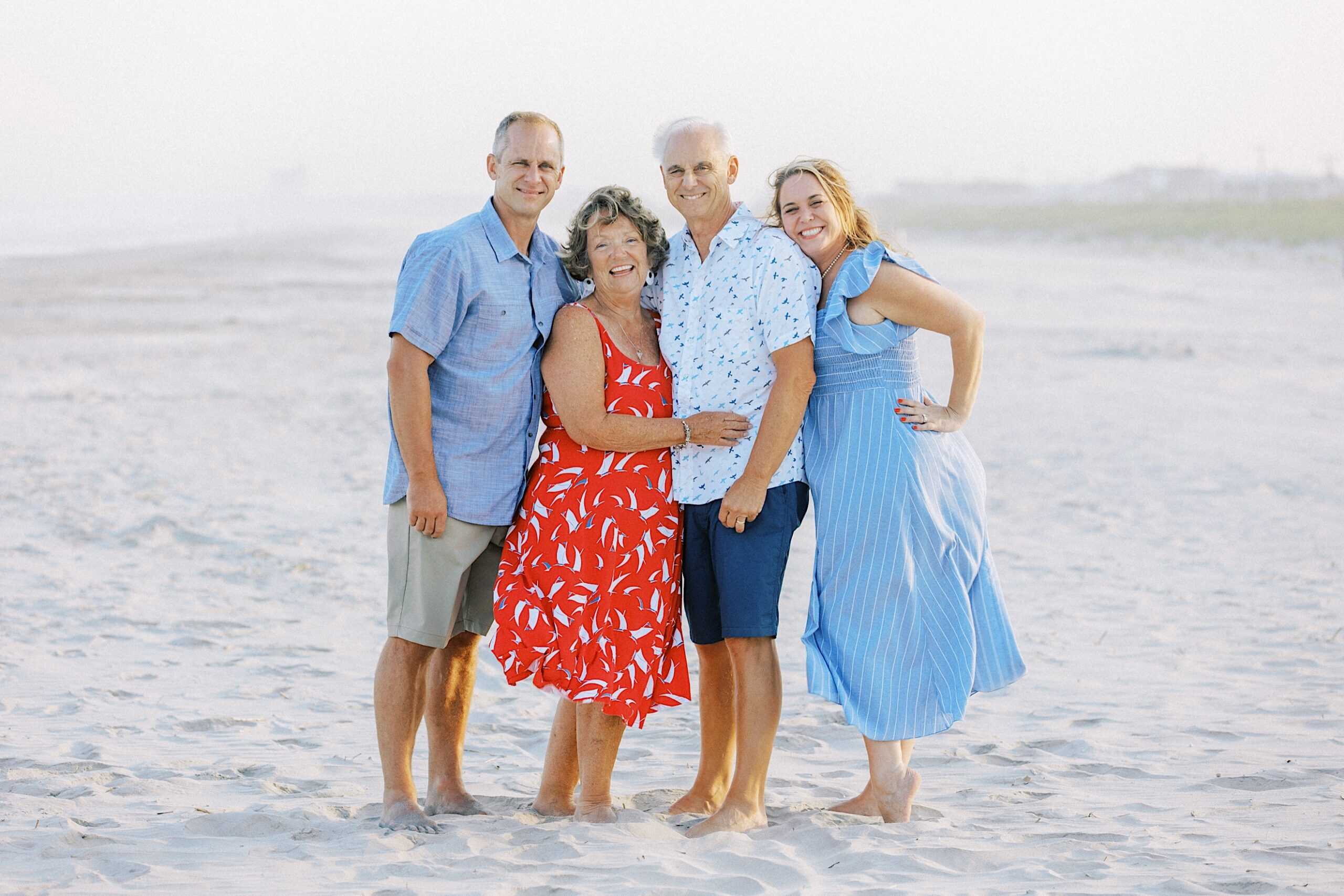 Four adults stand barefoot on a sandy beach, posing for Jersey Shore family photos. Two men wear blue shirts, one woman is in a red dress, and the other wears blue. The background is hazy, capturing a relaxed seaside moment.