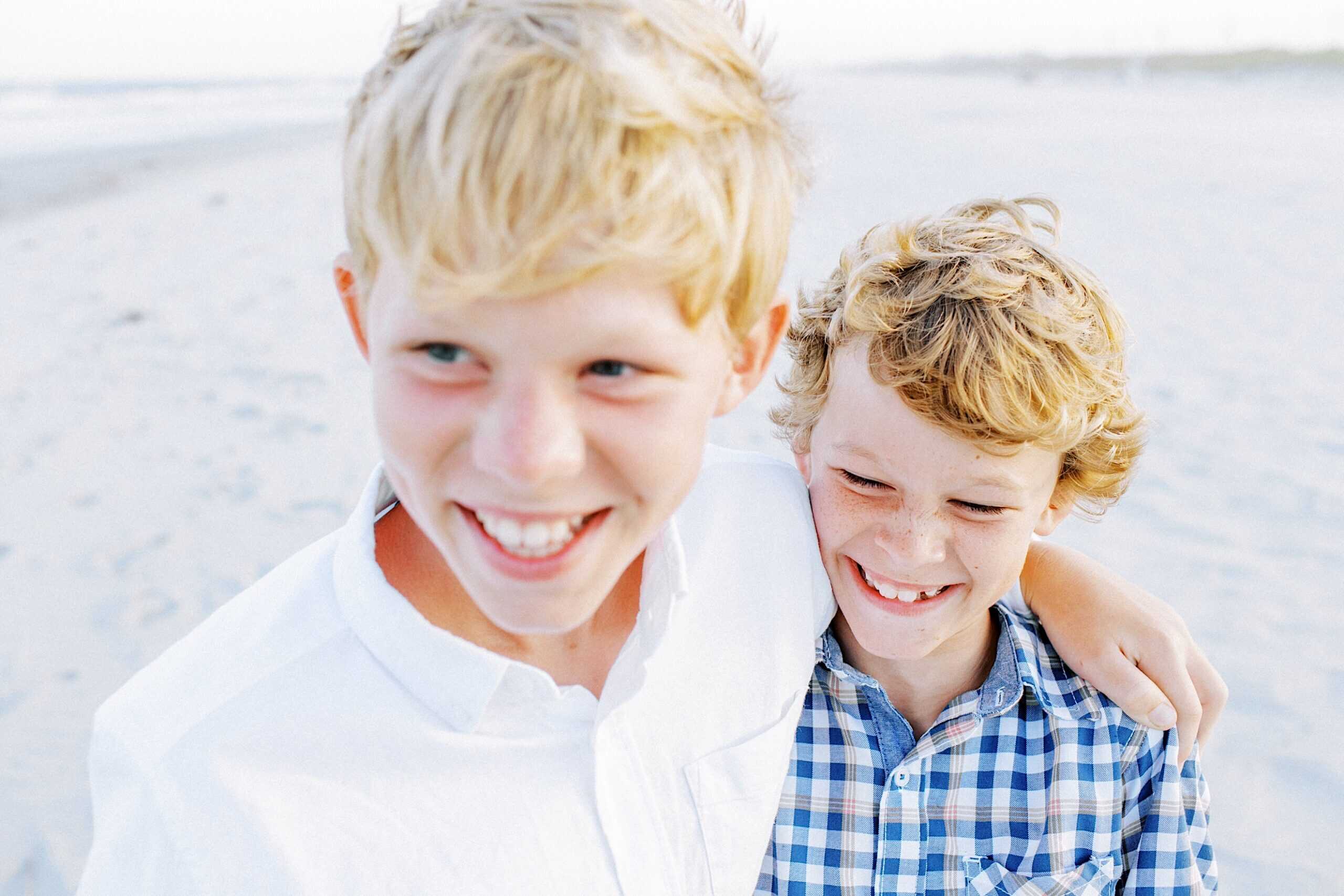 Two boys with blonde hair smile while standing close together on the sandy beach, perfect for Jersey Shore family photos. One wears a white shirt, the other sports a blue plaid shirt.