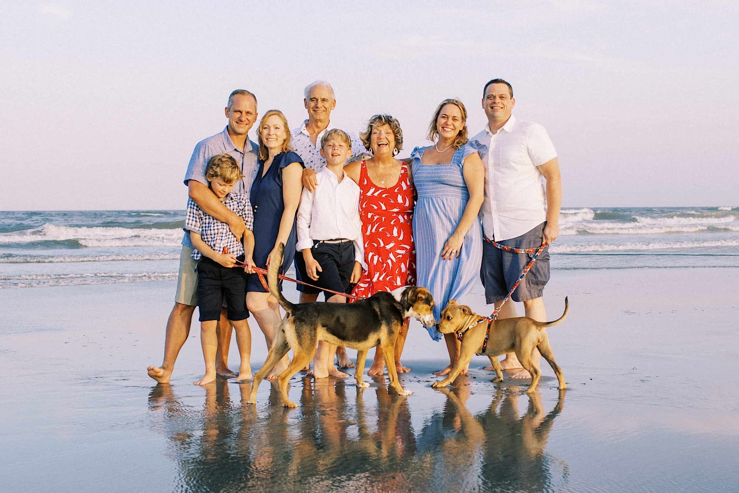 A group of eight people, including children, and two dogs stand together smiling on a beach with ocean waves in the background, capturing classic Jersey Shore Family Photos.