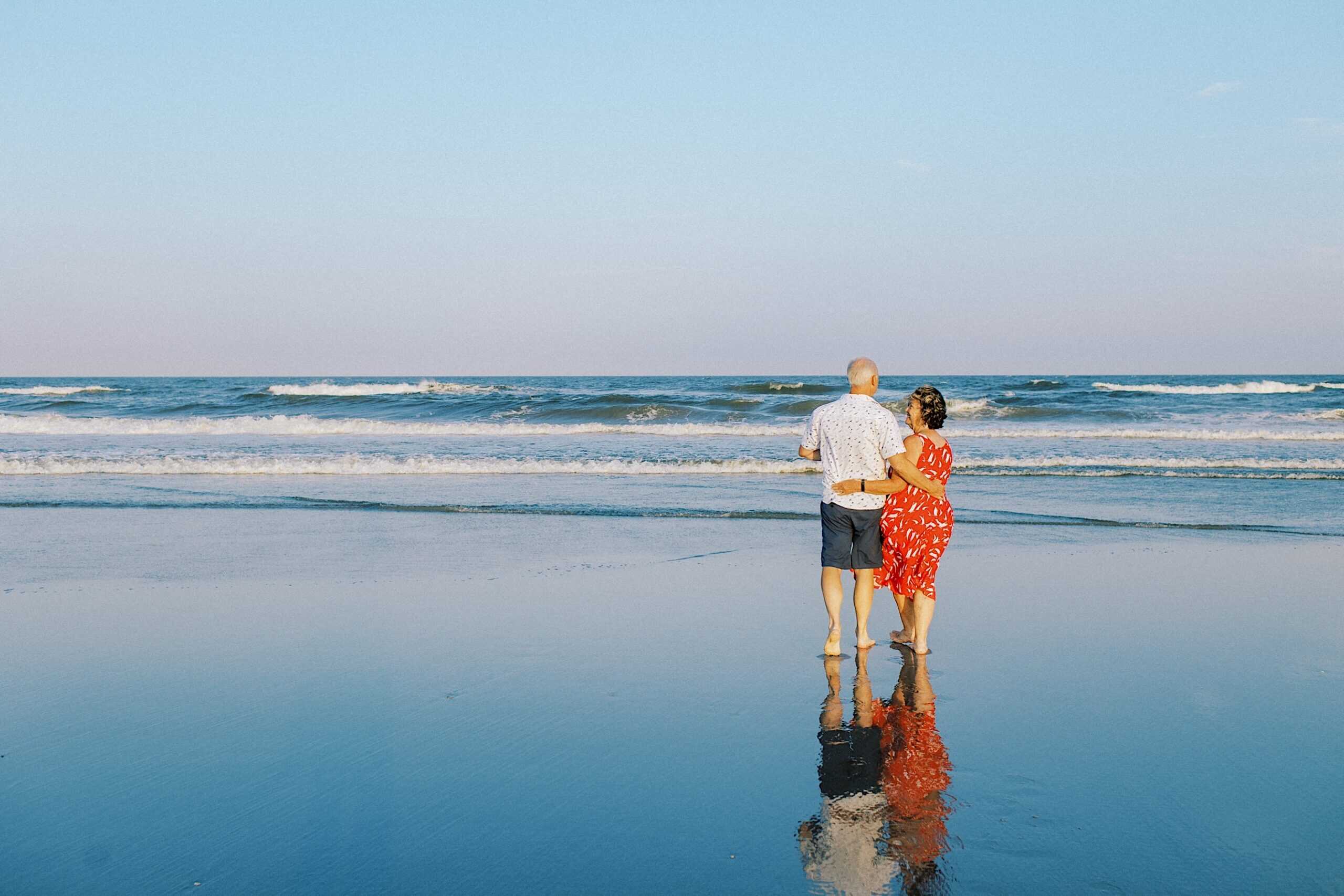 An older couple stands arm in arm on a Jersey Shore beach, facing the ocean, their reflection visible on the wet sand under a clear blue sky—a timeless moment perfect for family photos.