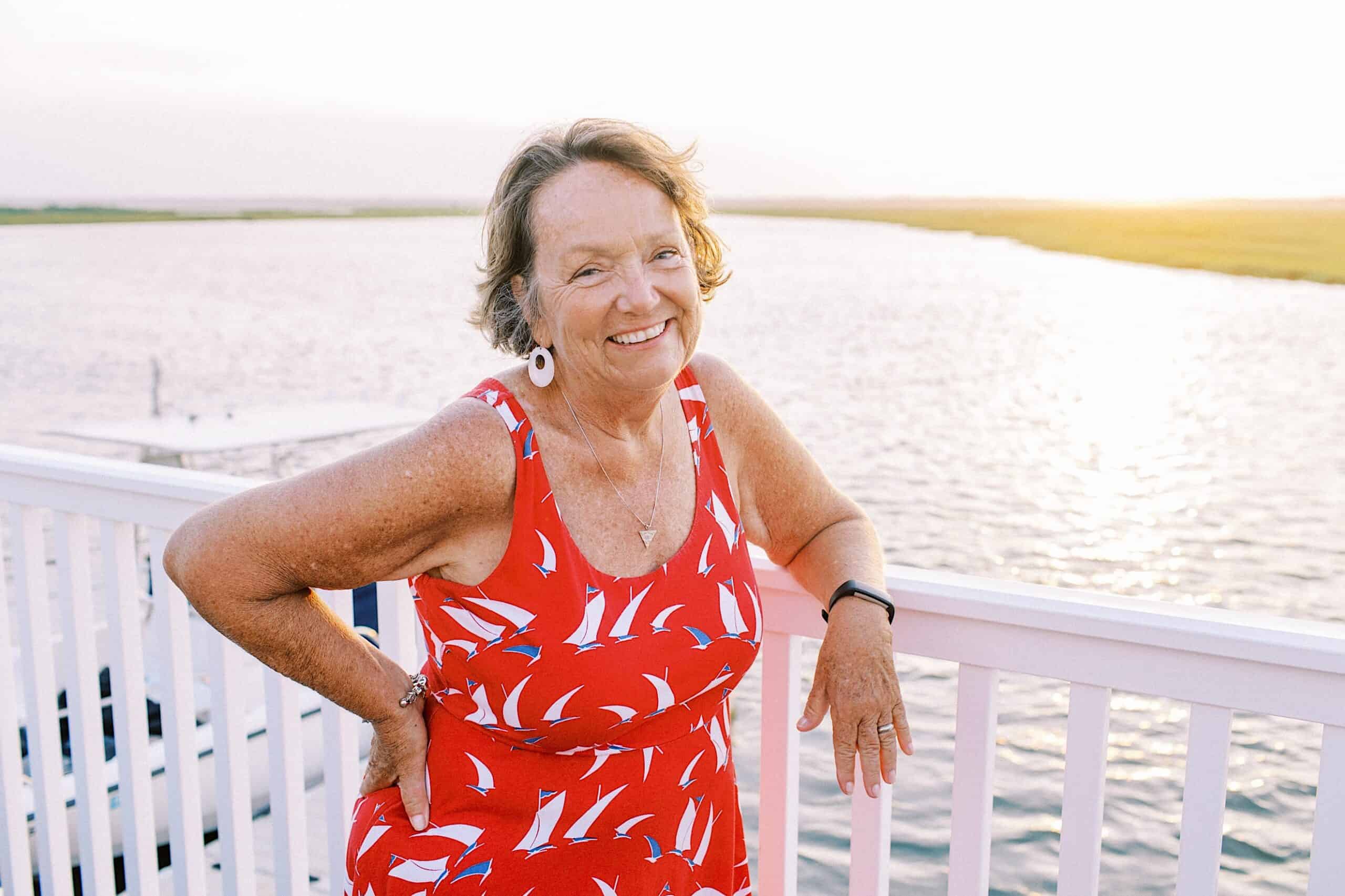 An older woman in a red dress with white sailboat patterns stands by a white railing at sunset, smiling at the camera—a timeless moment from our Jersey Shore Family Photos collection.