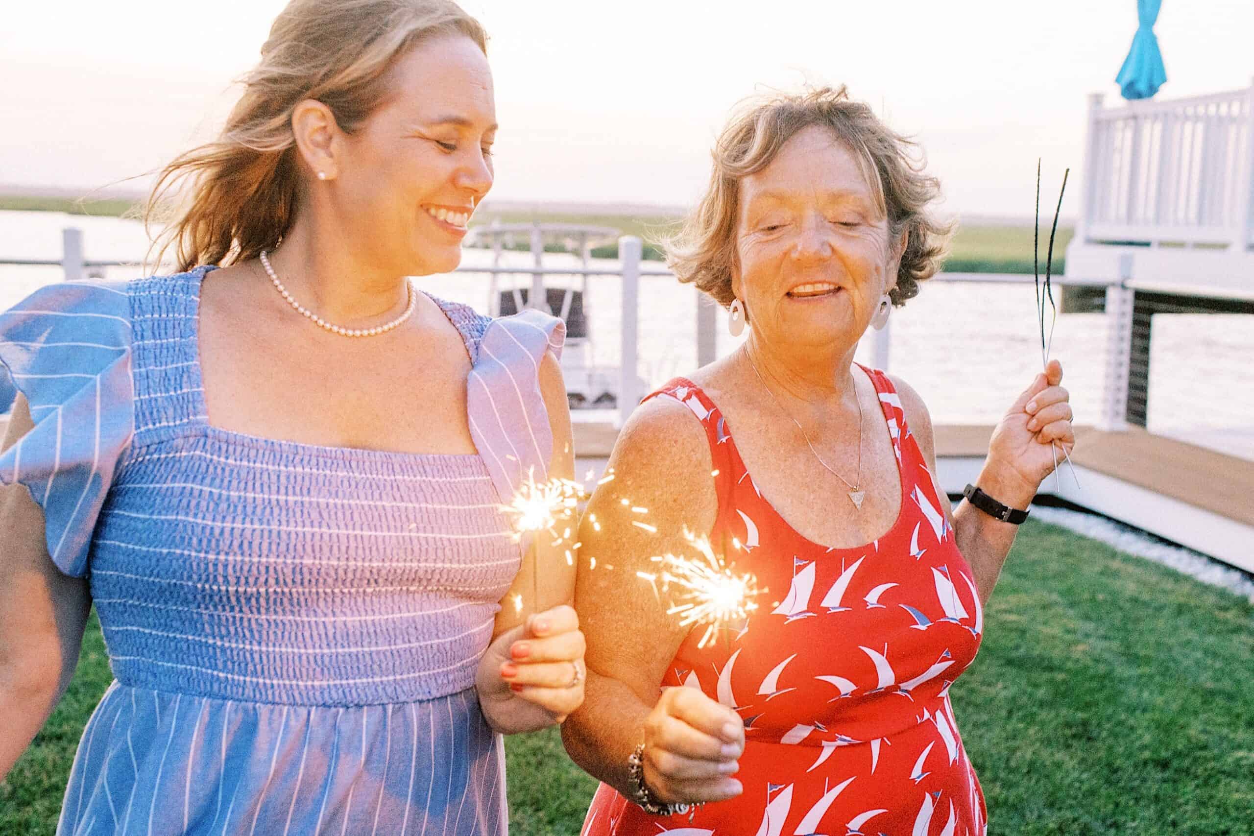 Two women standing outdoors near water, holding lit sparklers and smiling, capture a joyful moment reminiscent of Jersey Shore Family Photos, with a deck and grassy area in the background.
