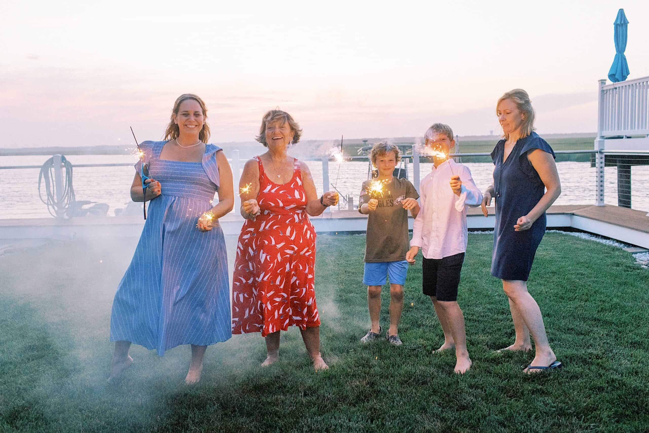 Five people stand barefoot on grass near water at sunset, holding lit sparklers and smiling, capturing the joy of Jersey Shore Family Photos.