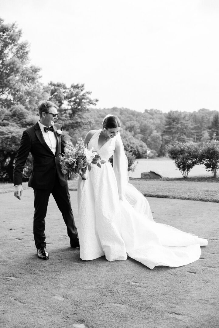 A bride and groom stand outdoors at an Overbrook Golf Club wedding. The bride, holding a bouquet, adjusts her dress while the groom stands beside her, both dressed formally. Vegetation and a body of water are visible in the background.