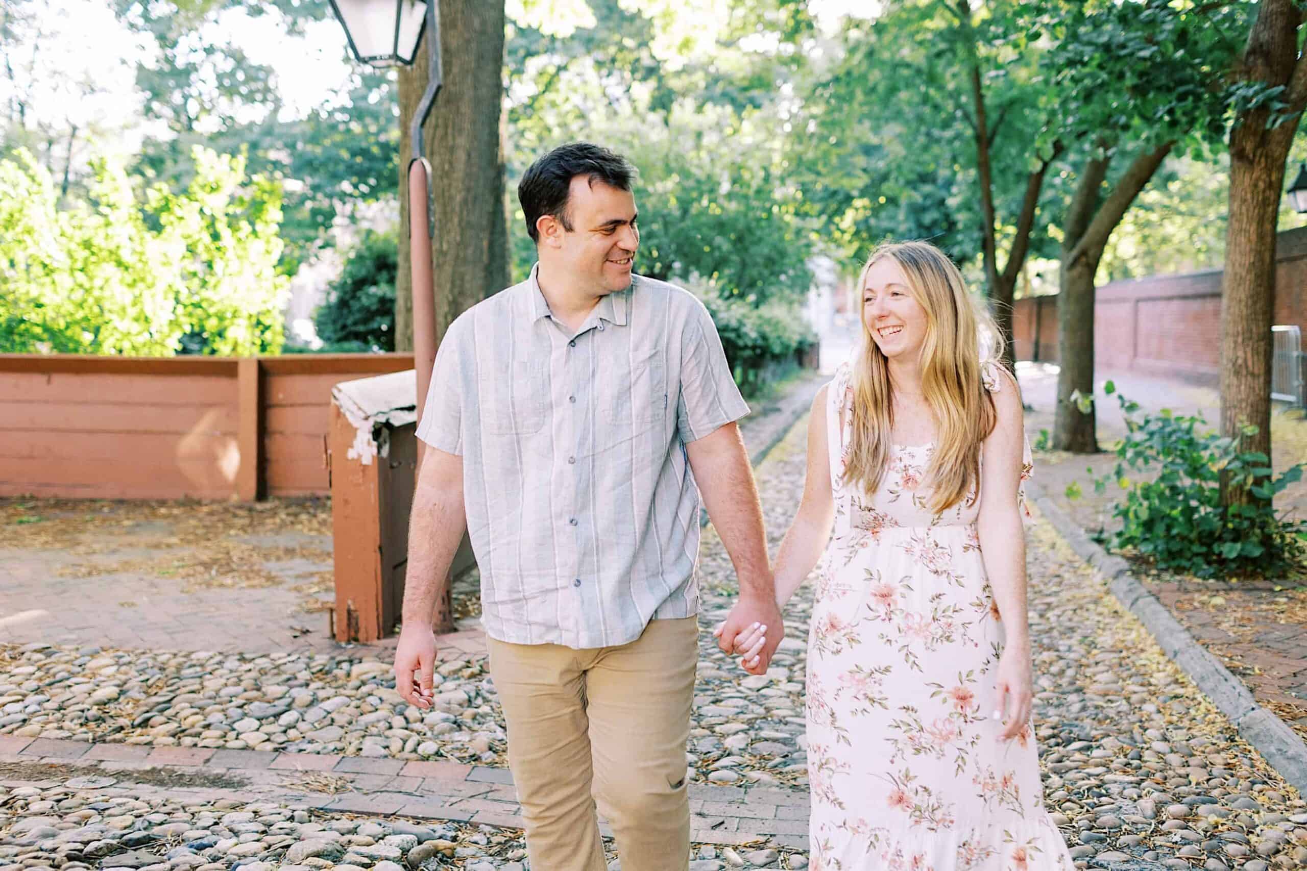 A man and woman walk hand in hand on a cobblestone path outdoors in Old City Philadelphia, surrounded by trees and wooden fences, both smiling—capturing timeless engagement photos.