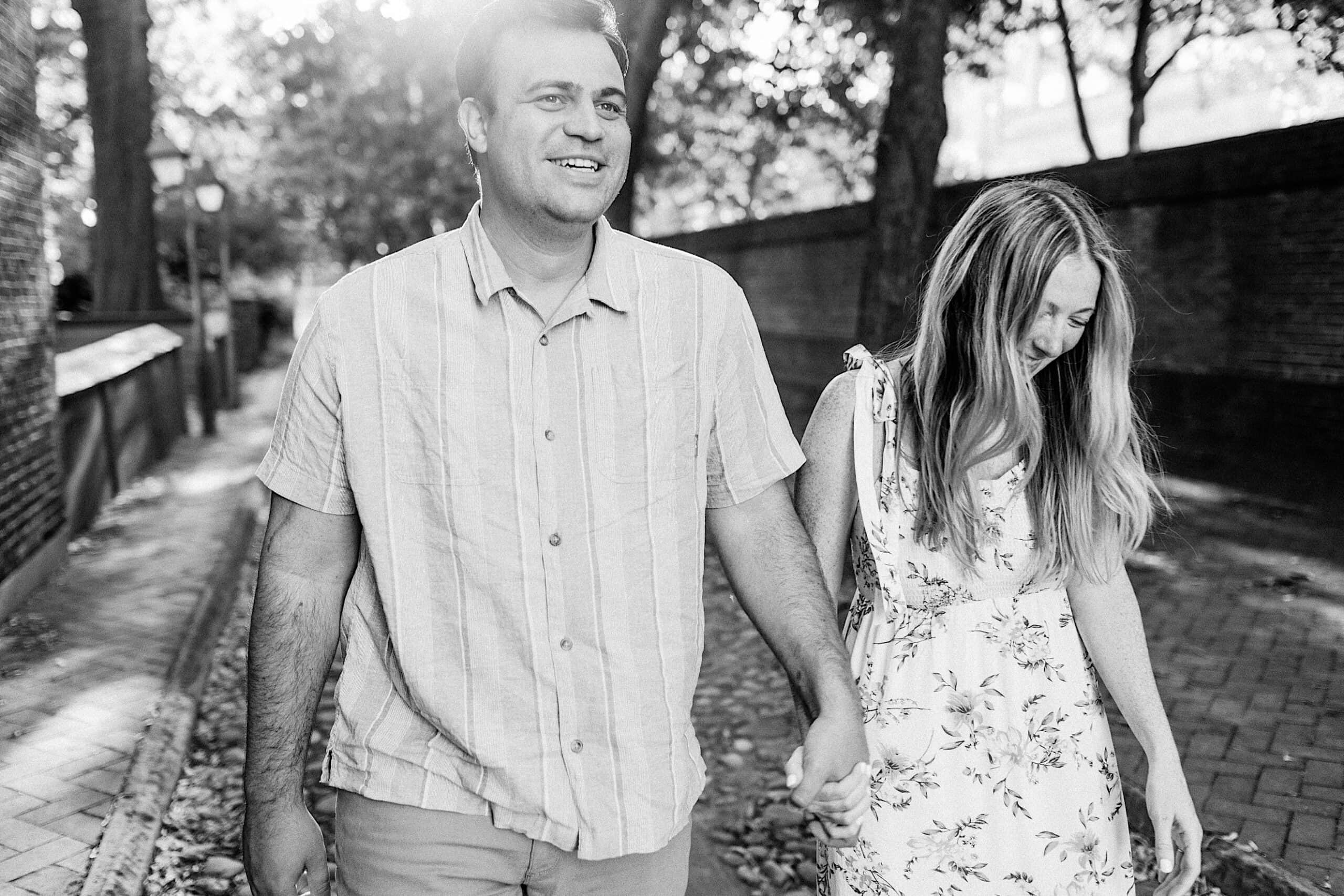 A man and woman walk hand in hand down a tree-lined brick path, both smiling; the photo is in black and white, capturing the timeless charm of Old City Philadelphia engagement photos.