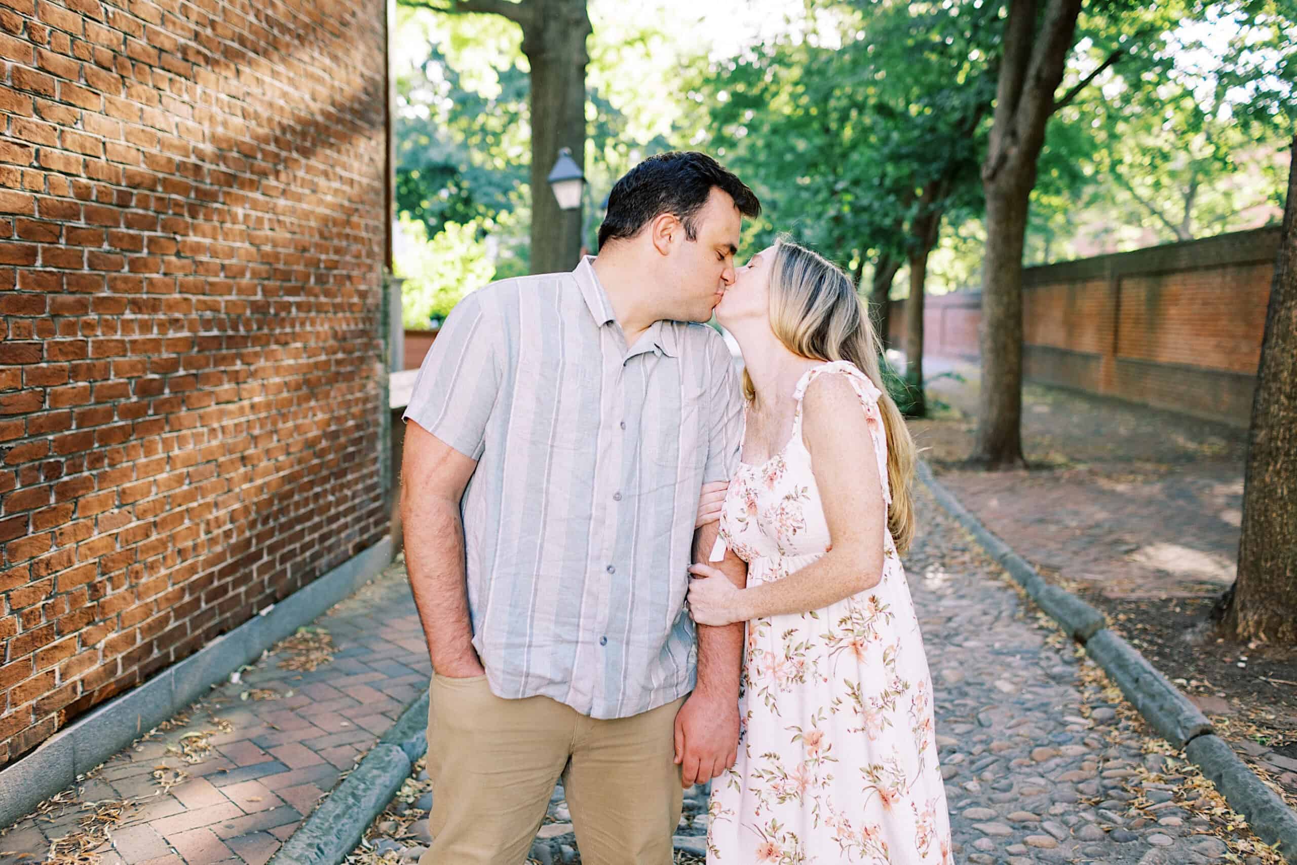 A couple stands on a cobblestone path by a brick wall, holding hands and kissing under trees on a sunny day—capturing romantic Old City Philadelphia engagement photos.