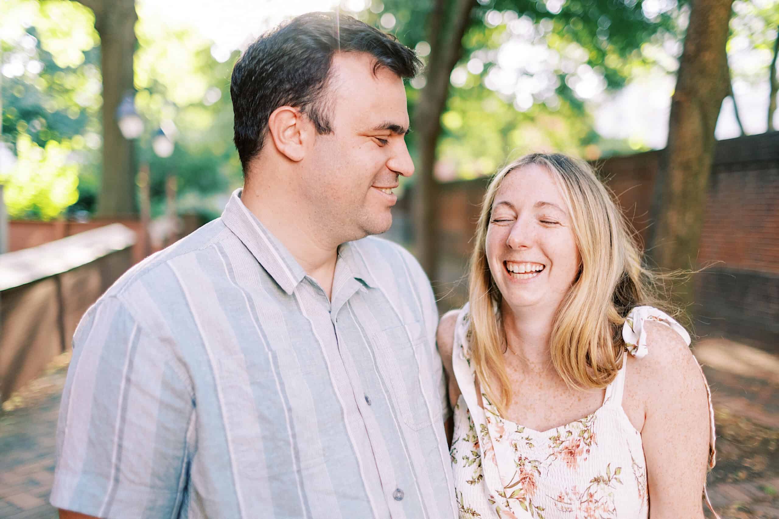 A man and woman stand outdoors on a sunny day in Old City Philadelphia, smiling and laughing together during their engagement photos. Trees and a brick wall are visible in the background.