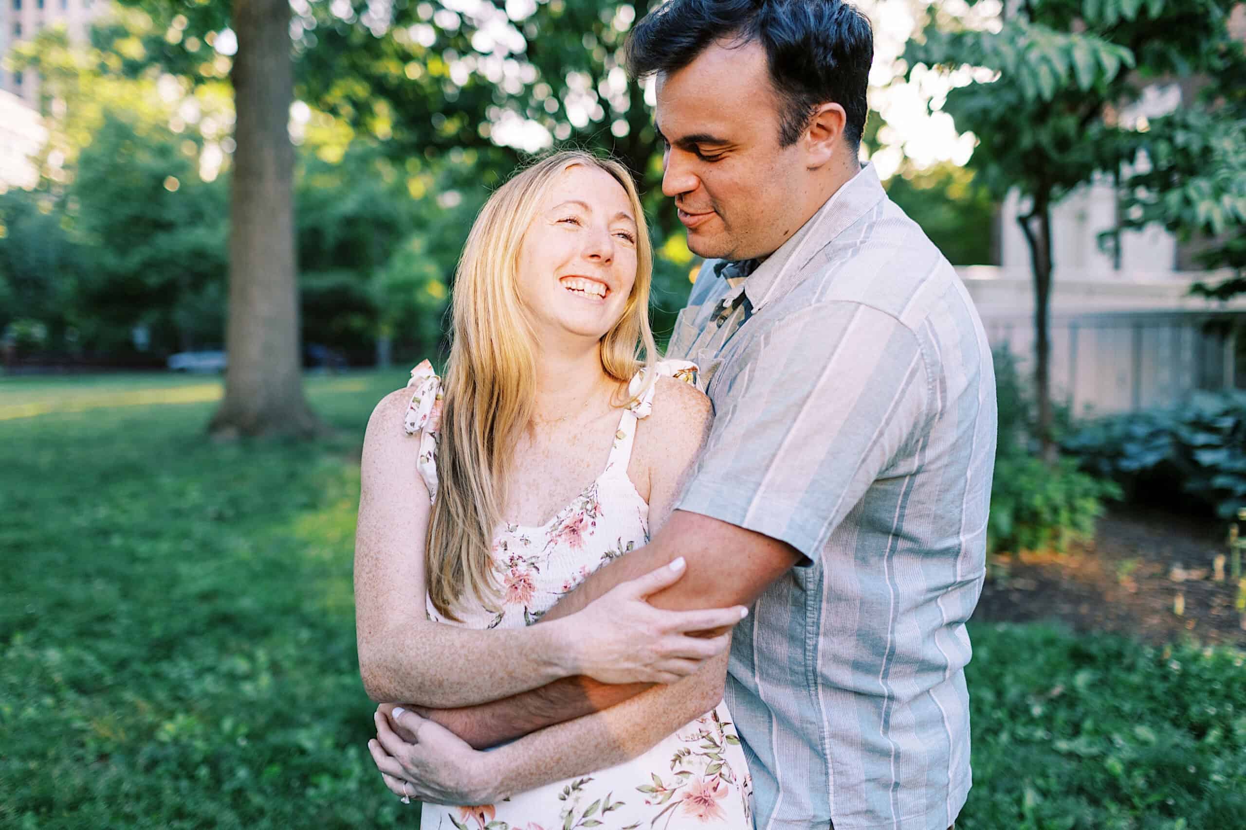 A man and woman stand close together in a park, smiling and embracing, surrounded by green trees and grass on a sunny day—capturing the charm of Old City Philadelphia engagement photos.
