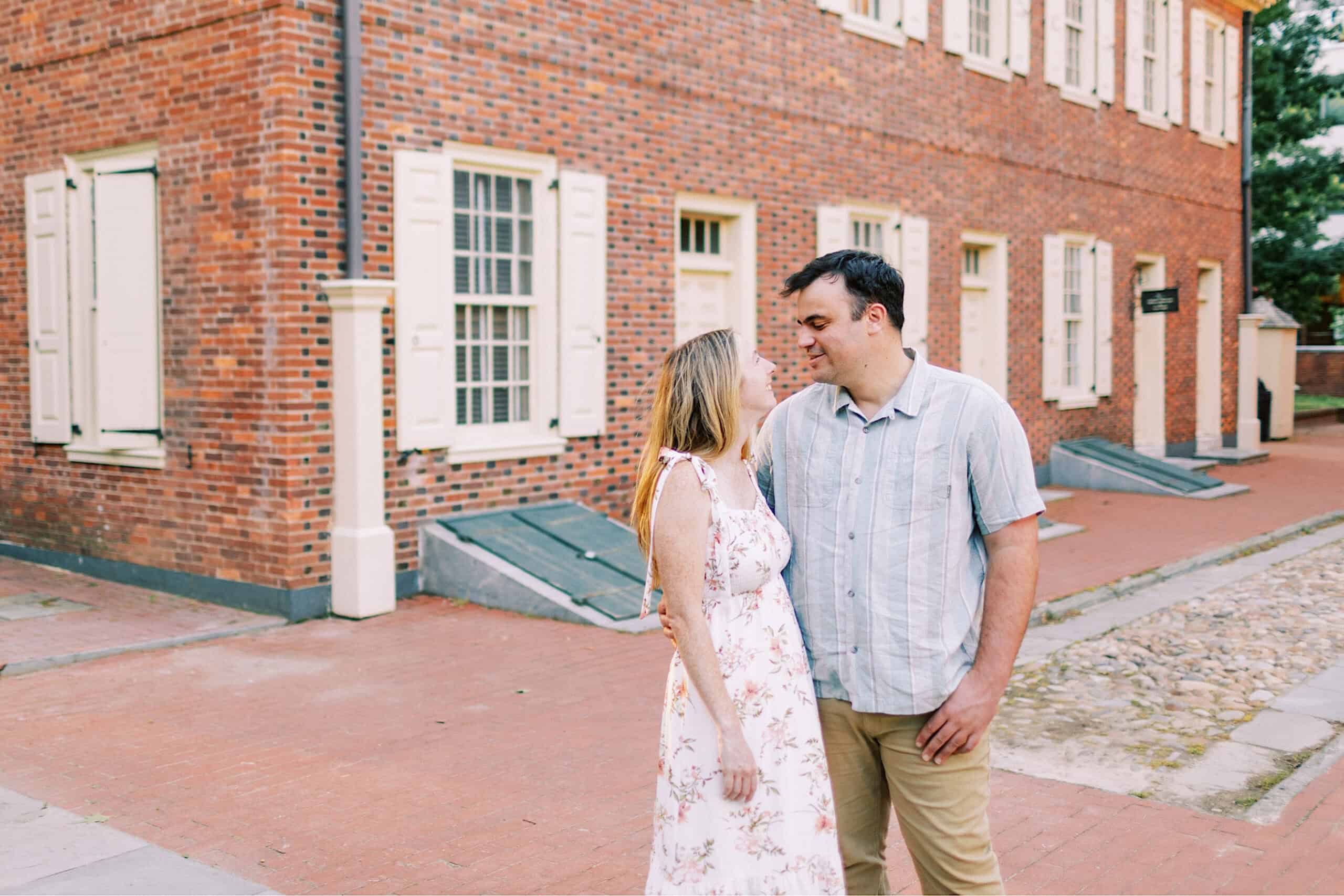 A couple stands close together on a brick sidewalk in front of a historic red brick building with white shutters, capturing timeless Old City Philadelphia engagement photos.