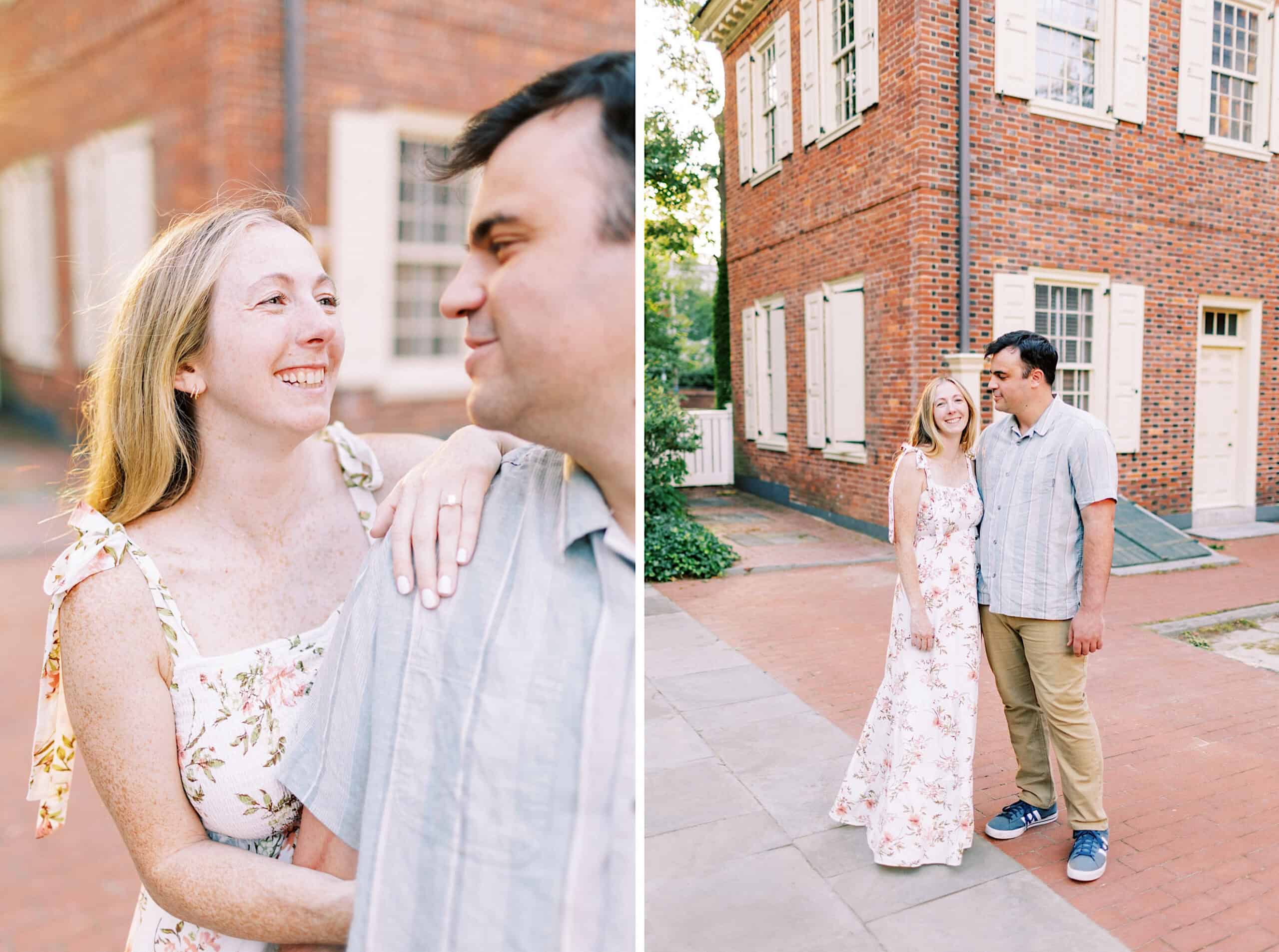 A woman in a floral dress and a man in a gray shirt stand together outside a red brick building, smiling at each other during their Old City Philadelphia engagement photos.