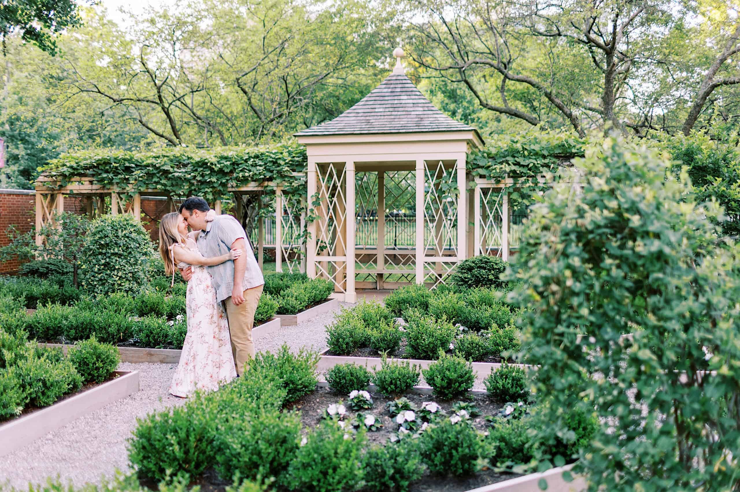 A couple stands in a garden, embracing and kissing near a small, open-sided gazebo surrounded by greenery and manicured bushes—capturing romantic Old City Philadelphia engagement photos.