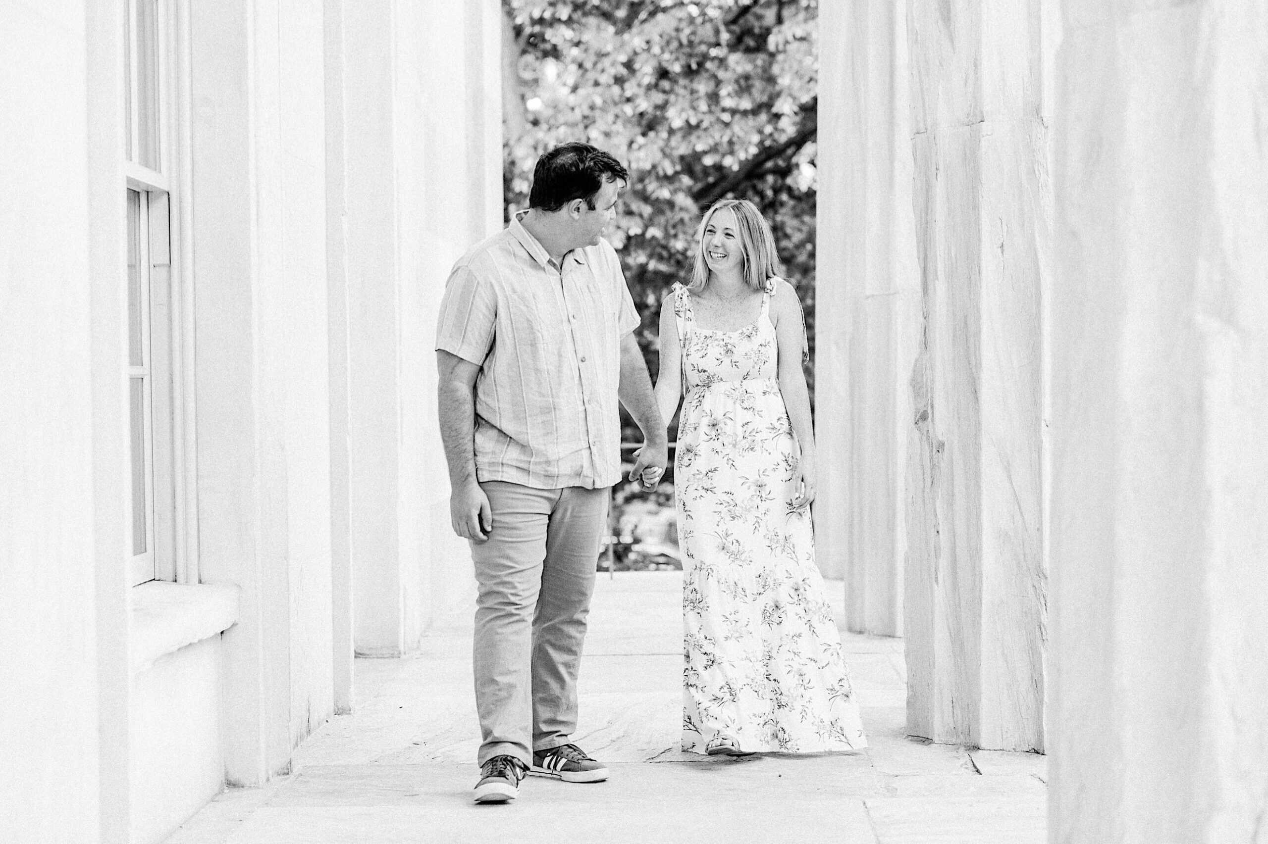 A man and woman walk hand in hand down a marble walkway, smiling at each other during their Old City Philadelphia engagement photos. The scene is outdoors with trees visible in the background.