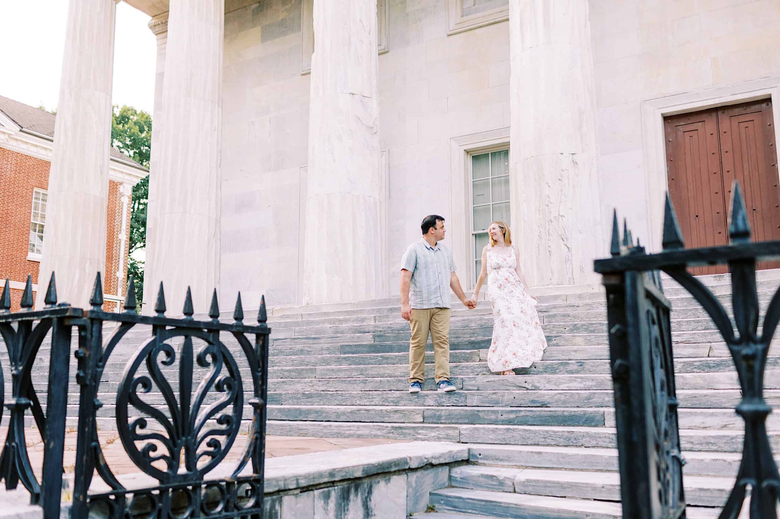 A man and woman hold hands while walking down stone steps outside a large building with tall white columns and decorative black iron railings, capturing timeless Old City Philadelphia engagement photos.
