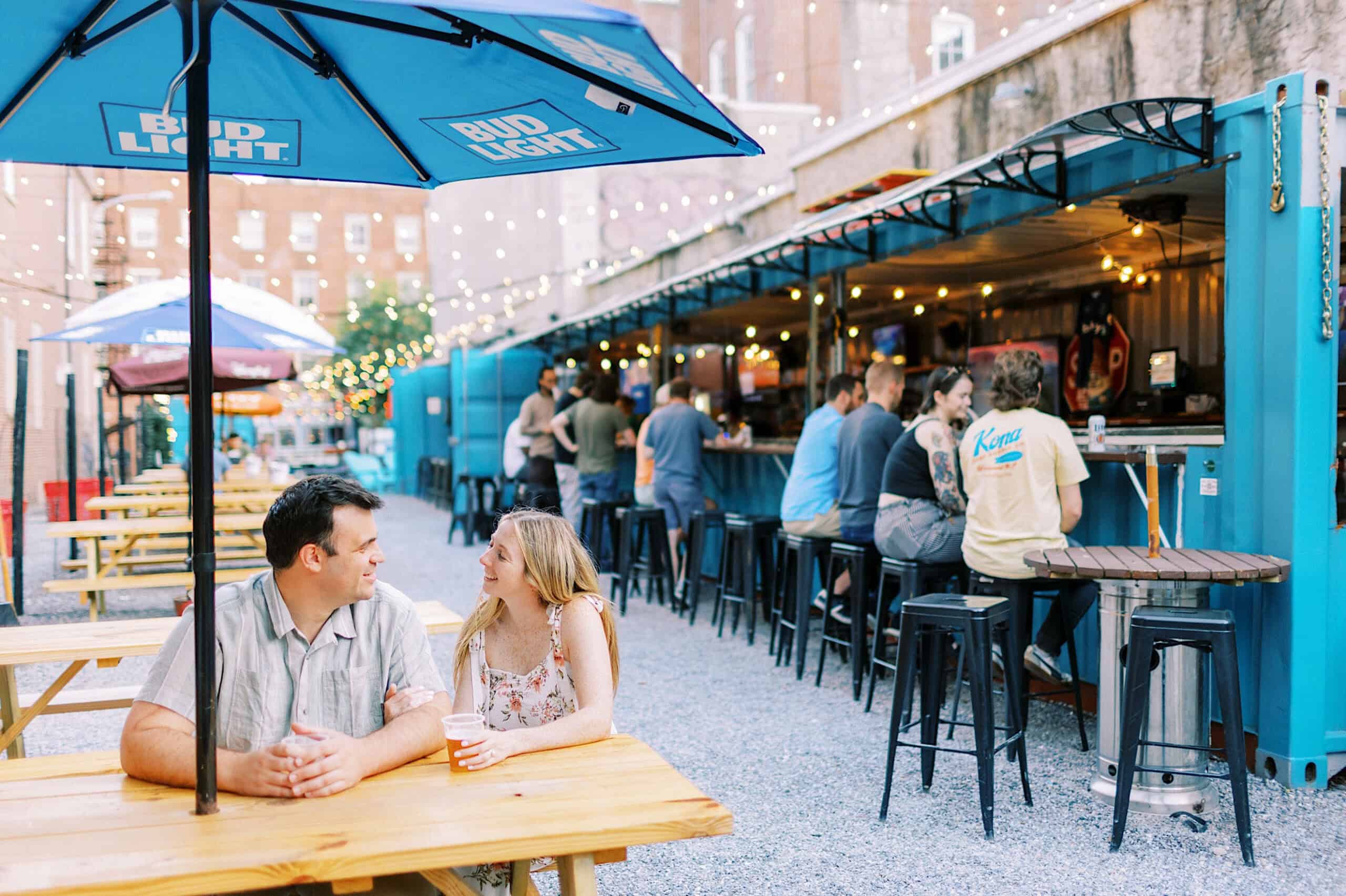 People sit at picnic tables and barstools in an outdoor bar area with blue shipping containers, string lights, Bud Light umbrellas—an inviting spot that’s perfect for casual Old City Philadelphia engagement photos.