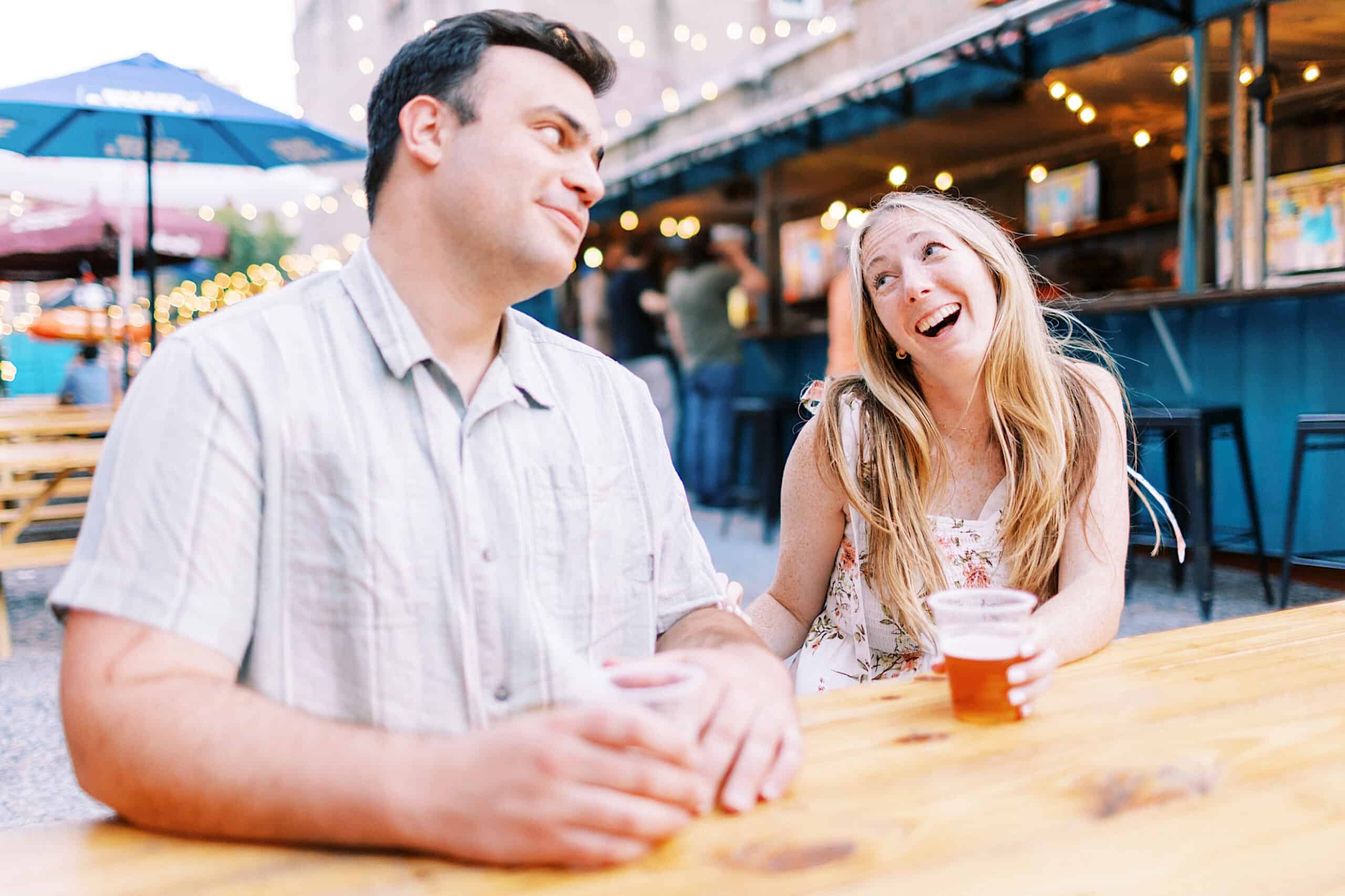 A man and woman sit at an outdoor table with drinks, smiling and laughing under string lights, surrounded by people—capturing candid Old City Philadelphia engagement photos.