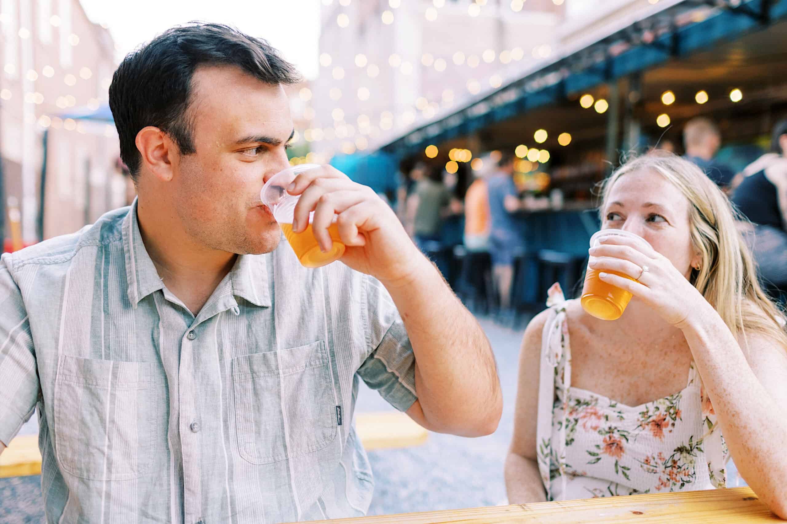 A man and a woman sit at an outdoor table in Old City Philadelphia, drinking beer from plastic cups, with string lights and other people in the background—perfect for candid engagement photos.