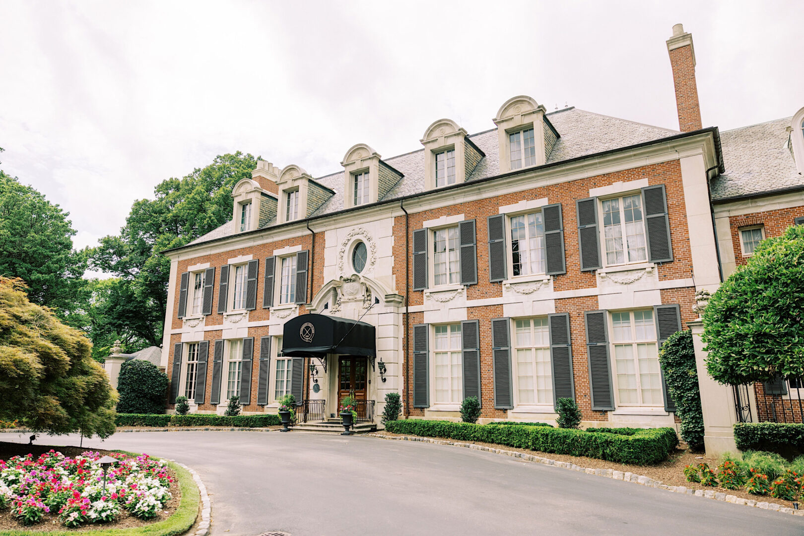 A large, elegant brick building with white trim, multiple windows, and a black arched canopy over the entrance, surrounded by manicured bushes and a floral garden; the perfect setting for an Overbrook Golf Club Wedding in Villanova, just minutes from Philadelphia.