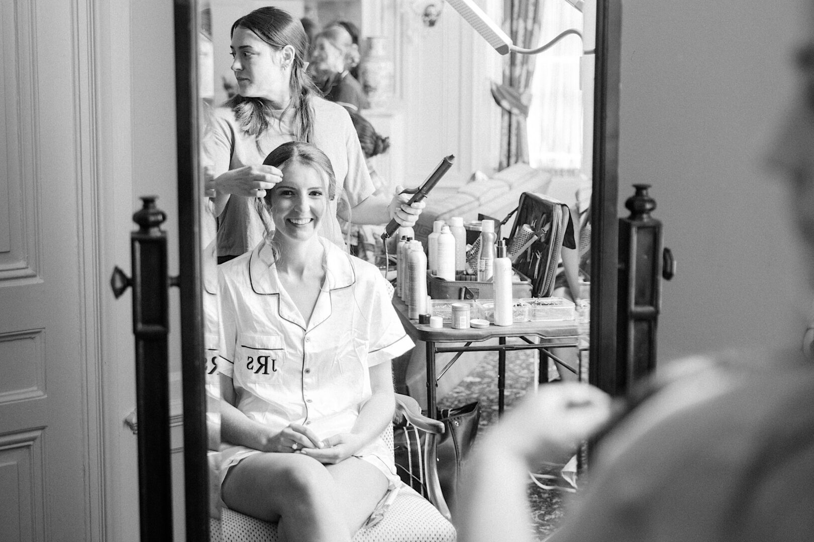 A woman in a robe labeled "Mrs." smiles in a mirror while getting her hair styled by another woman, preparing for her Overbrook Golf Club wedding. Various hair styling tools and products are visible on a table in the background, capturing the excitement of this special Philadelphia event.