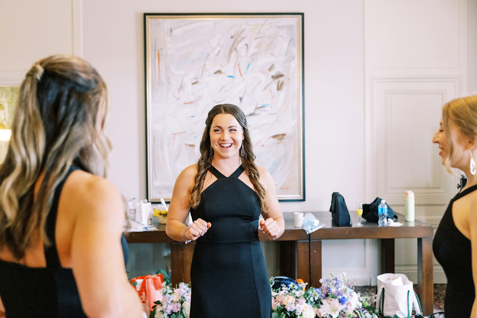 Three people stand in a room, smiling and conversing at what appears to be an elegant Overbrook Golf Club wedding. A painting hangs on the wall in the background. The table behind them holds various items, including a candle and flower arrangements, adding charm to this lovely Philadelphia setting.