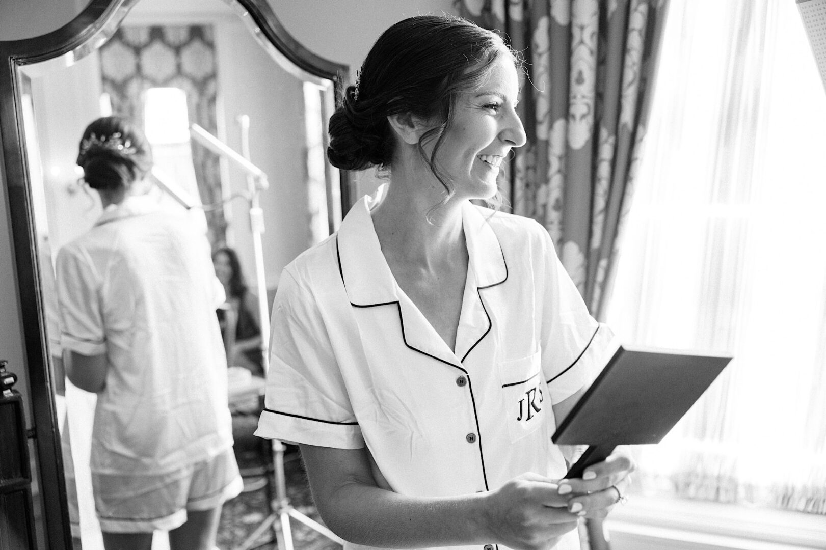 A woman in matching robe and shorts stands in front of a mirror, holding a smaller hand mirror, smiling. The room, adorned with patterned wallpaper and curtains, is bathed in natural light from the window—a perfect start to her day before attending an Overbrook Golf Club Wedding.