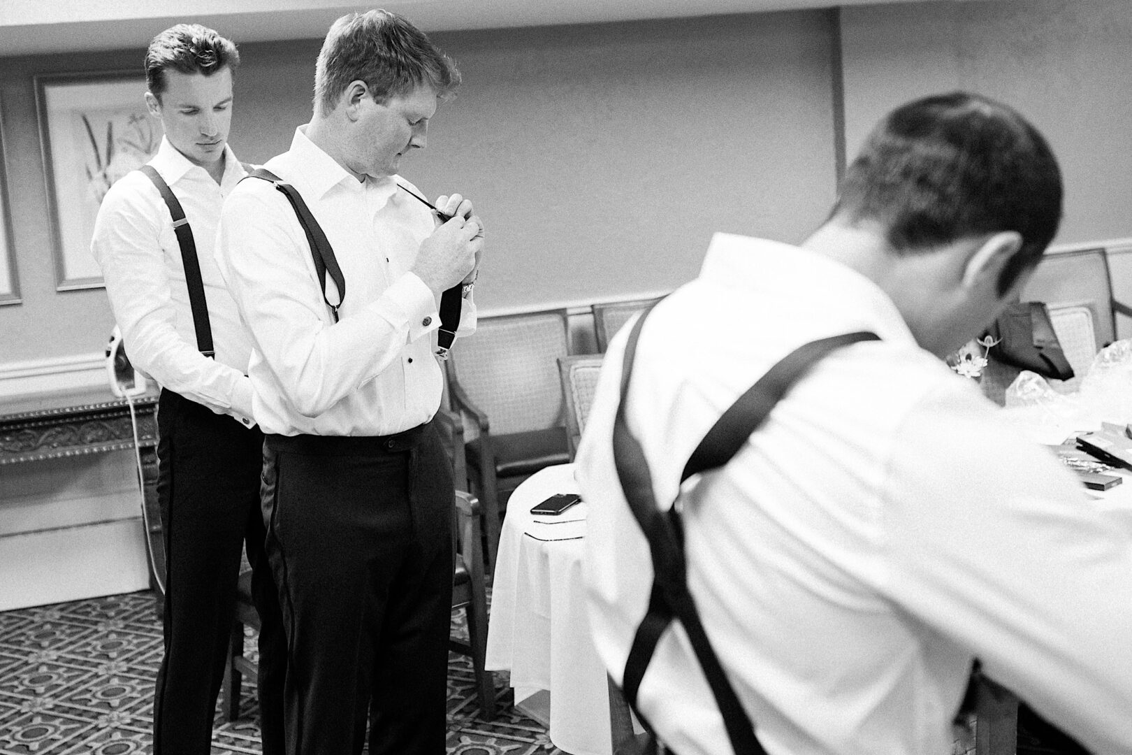 Three men wearing suspenders adjust their clothing in a room with chairs and a table, preparing for an Overbrook Golf Club wedding in Philadelphia.