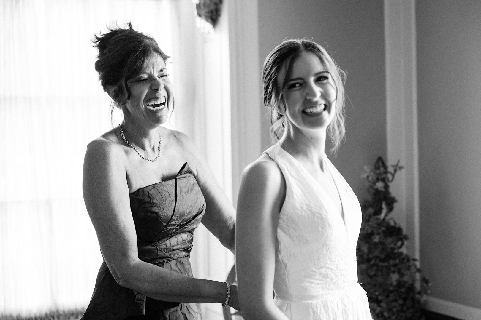 A woman in a formal dress smiles and adjusts the back of another woman's sleeveless dress in a well-lit room at an Overbrook Golf Club wedding. Both women, likely from Philadelphia, appear happy.