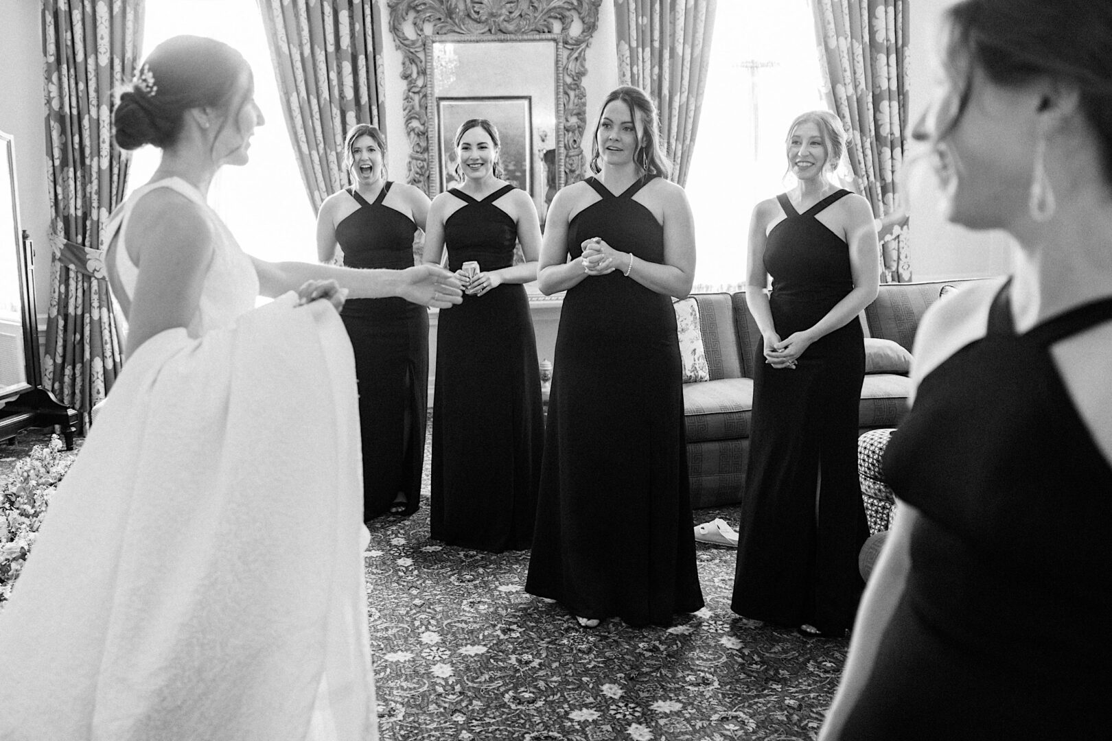 At an Overbrook Golf Club wedding in Philadelphia, a bride in a white dress interacts with four women in black gowns, all standing in a decorated room with curtains and a large mirror.