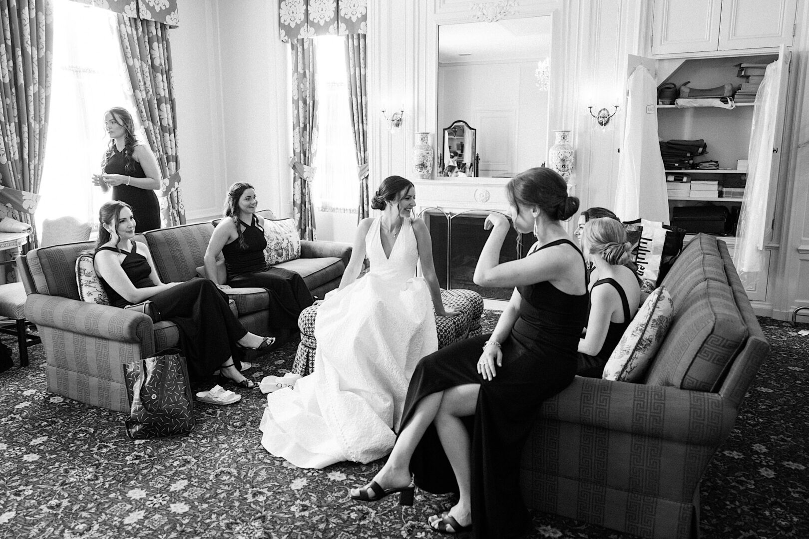 Black and white photo of a bride in a white gown seated on a couch, surrounded by five bridesmaids in black dresses, in a room with large windows and ornate curtains at the Overbrook Golf Club Wedding near Villanova, Philadelphia.