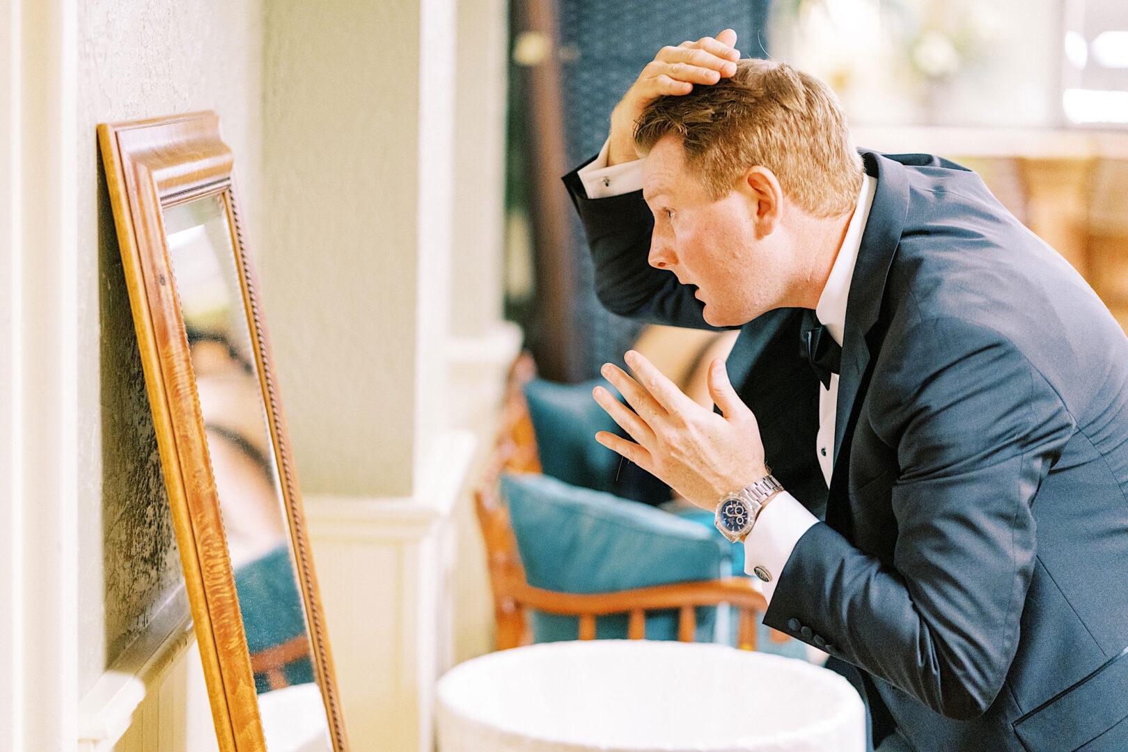 Man in a tuxedo adjusts his hair while looking into a mirror, preparing for an Overbrook Golf Club Wedding in Philadelphia. The well-decorated room adds to the anticipation of the formal event.