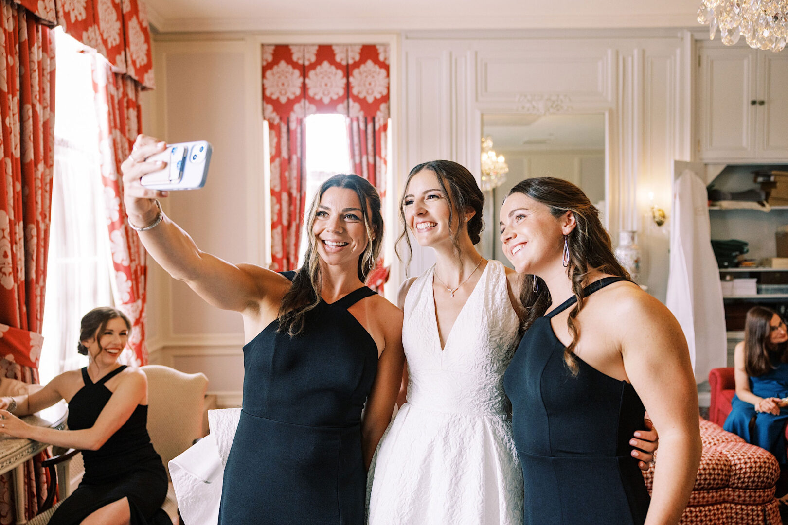 A bride and two bridesmaids pose for a selfie, while others sit in the background of an elegantly decorated room at the Overbrook Golf Club Wedding near Villanova.