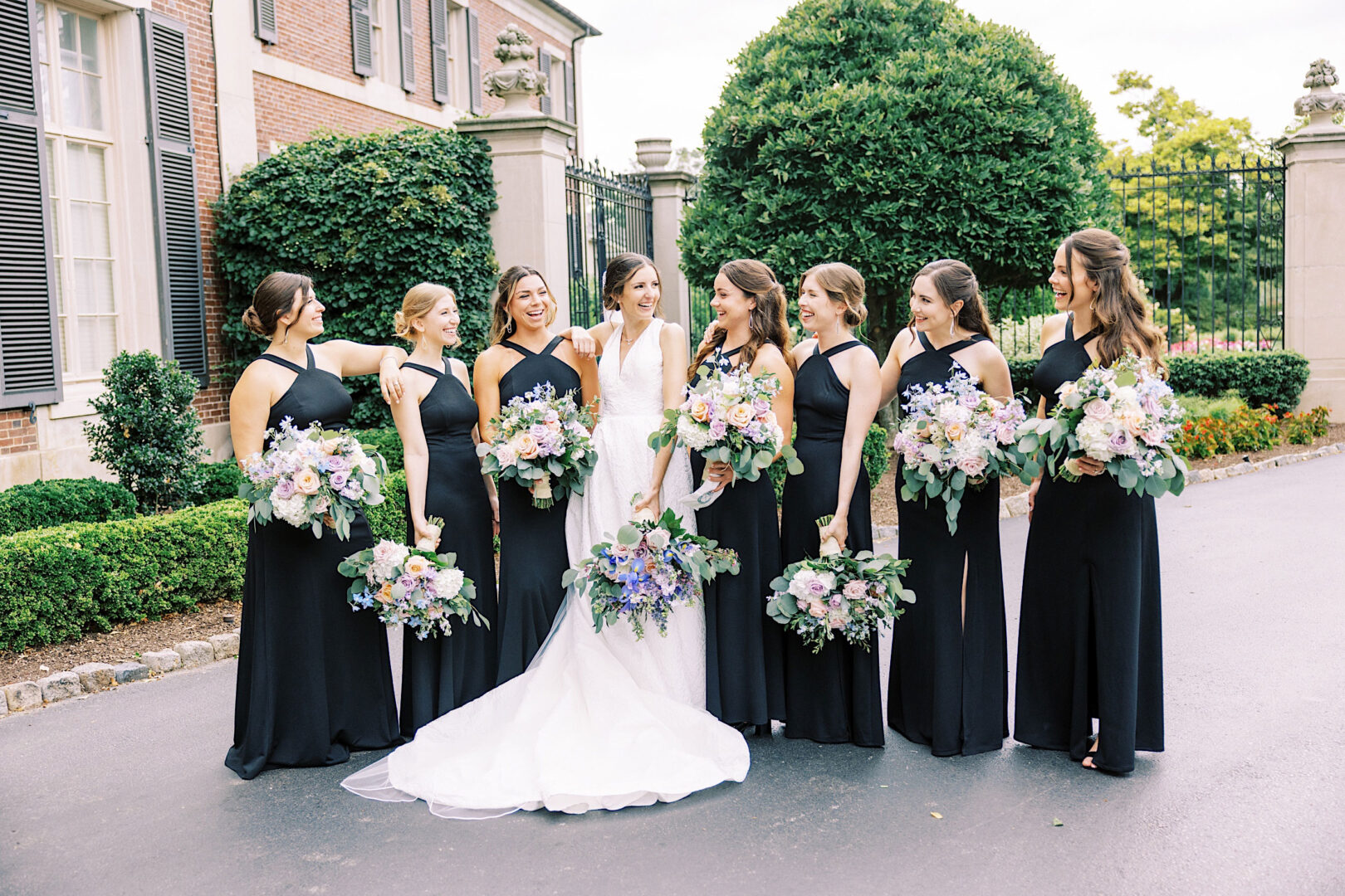 A bride in a white gown stands with six bridesmaids in black dresses, all holding bouquets, outside a building with large bushes and an ornate iron fence in the background at an elegant Overbrook Golf Club wedding near Philadelphia.