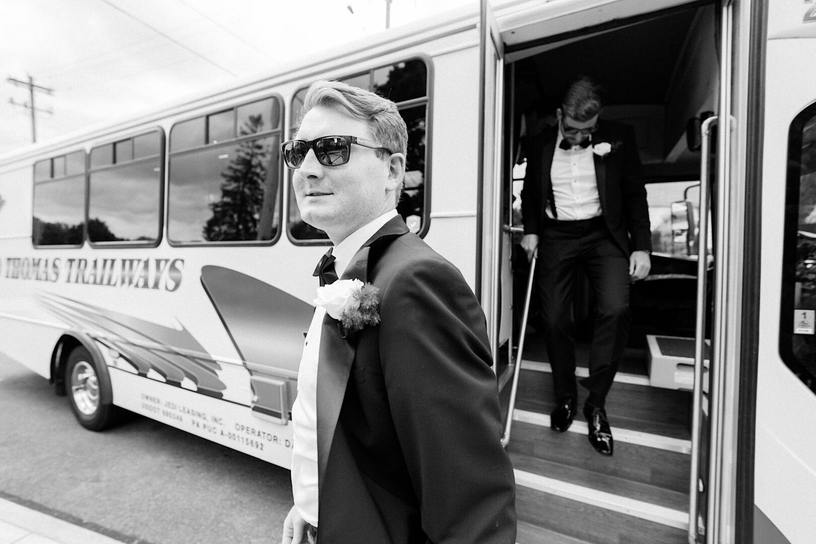 Two men in tuxedos exit a "Thomas Trailways" bus at the Overbrook Golf Club in Villanova. The man in front wears sunglasses and a boutonniere, while the other descends the steps behind him. The scene occurs in daylight, setting a perfect backdrop for this Philadelphia wedding.