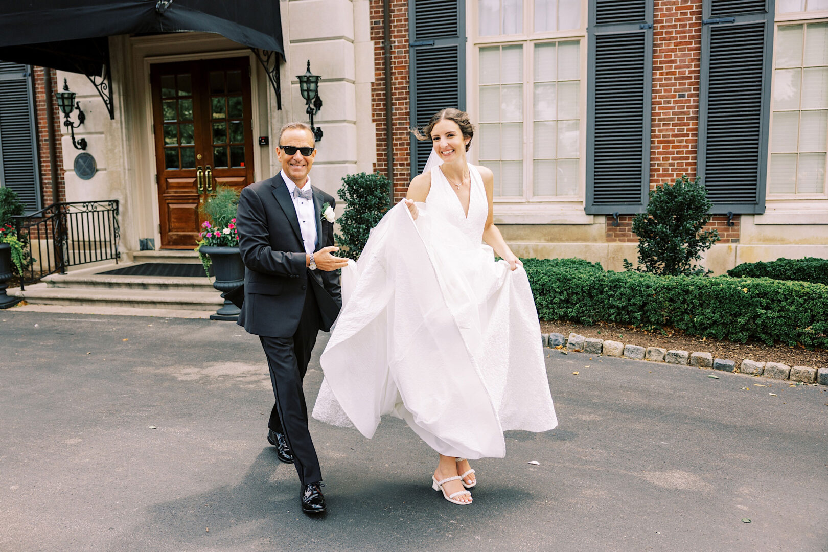 A bride and an older man, both smiling and dressed formally, walk arm-in-arm outside a brick building with black shutters and a wooden door. The bride, holding up her white gown to walk, radiates joy as they celebrate their Overbrook Golf Club Wedding near Villanova.