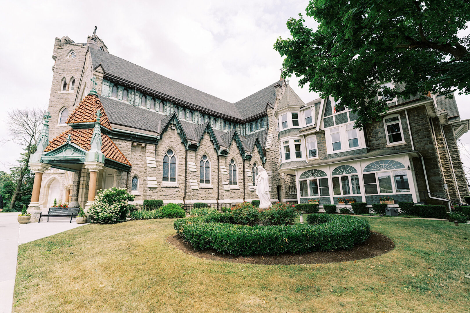 A stone church with a steep roof is adjacent to a large house with arched windows, reminiscent of Villanova's classic architecture. A neatly trimmed garden and statue are in the foreground.