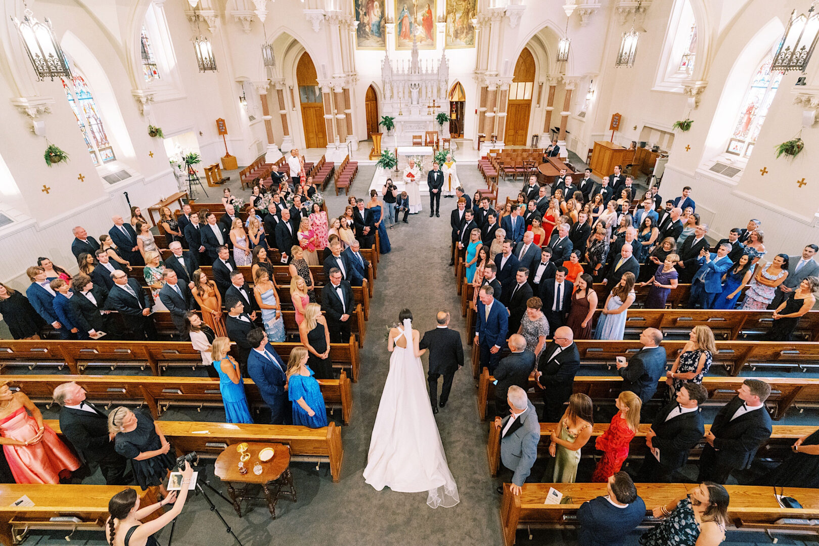 A bride walks down the aisle escorted by a man in a church filled with seated guests on either side, all facing towards the altar where the groom and officiant stand waiting, setting a perfect beginning to their Overbrook Golf Club Wedding celebration.