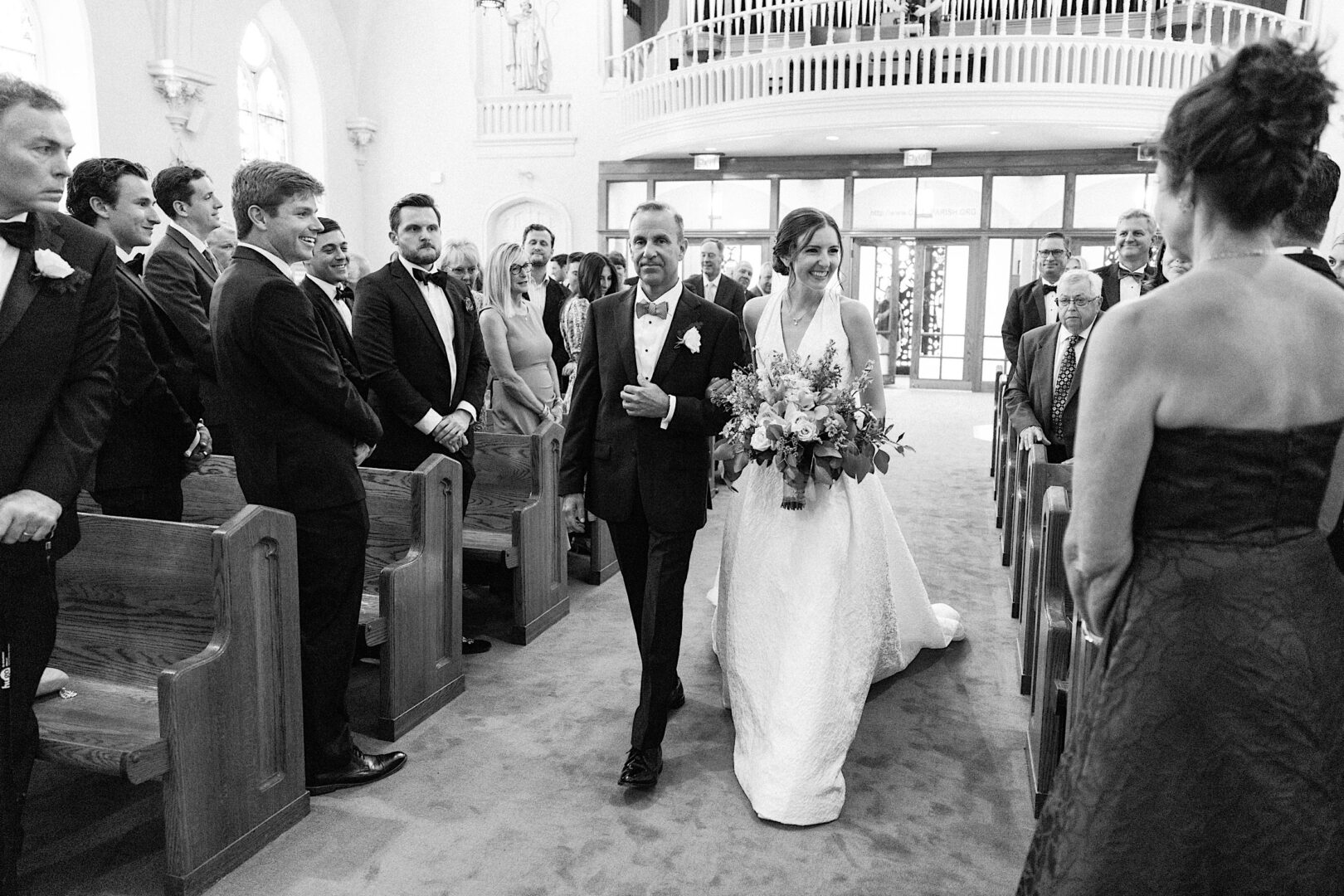 A bride walks down the aisle with an older man in a suit at a Villanova church. Guests stand on either side, some looking towards the bride, others towards the camera. The bride, set against this picturesque Philadelphia backdrop, carries a bouquet.