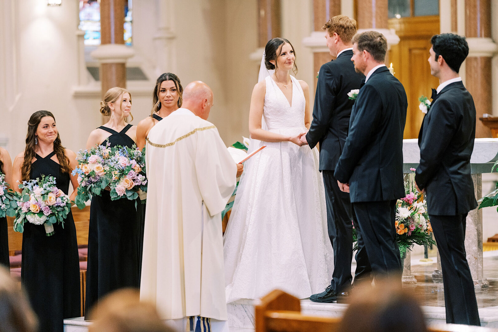 A bride and groom stand at the altar during their wedding ceremony at Overbrook Golf Club, facing a clergyman. Bridesmaids hold bouquets while groomsmen stand nearby. The setting is exquisite, with lush greenery providing a beautiful backdrop instead of traditional stained glass windows.