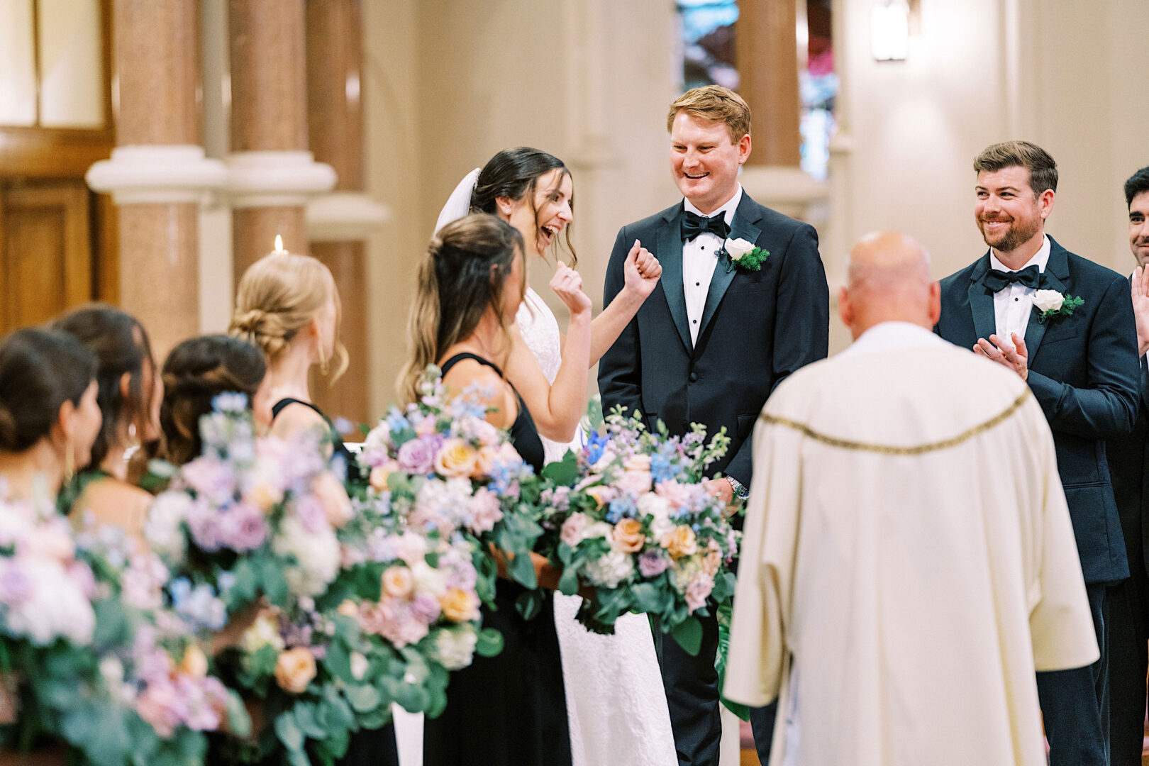 A bride and groom stand at the altar during an Overbrook Golf Club Wedding in Villanova, surrounded by wedding party members holding floral bouquets. The scene features a priest and ornate architectural details in the background, capturing the elegance of a Philadelphia celebration.