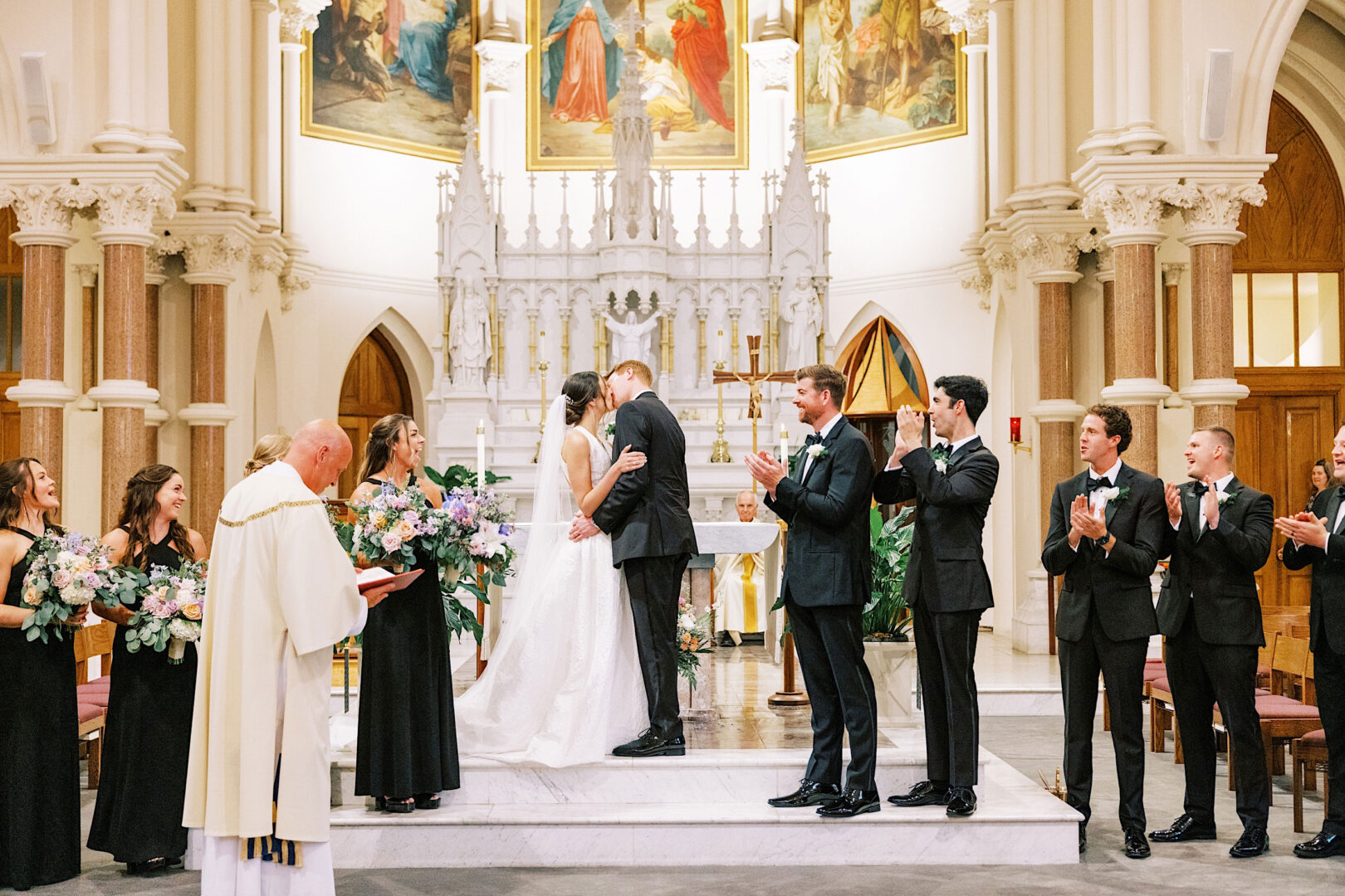 A bride and groom share a kiss at the altar during their wedding ceremony at Overbrook Golf Club in Villanova, surrounded by their wedding party and officiated by a priest.