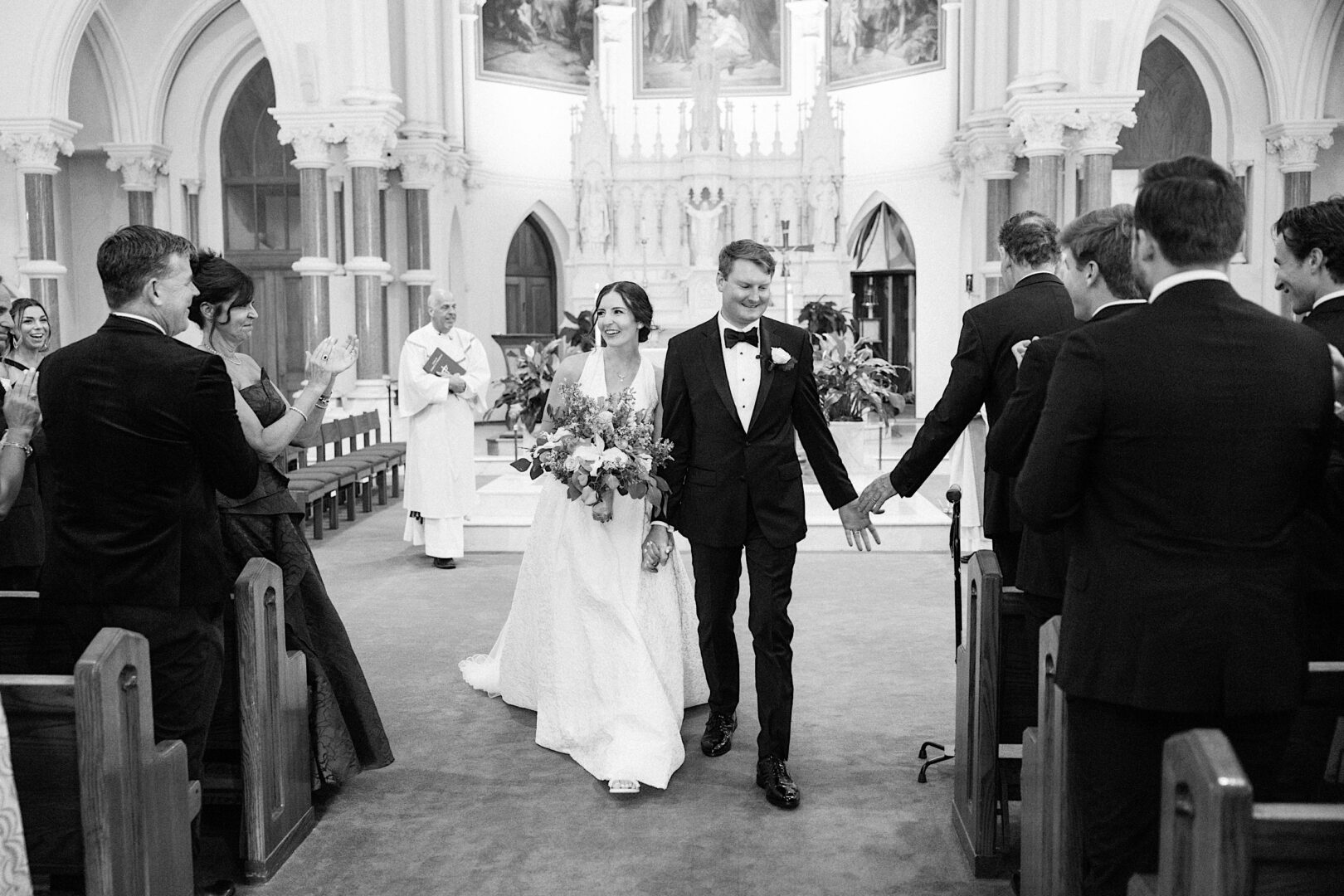 A newlywed couple walks down the aisle in a church, holding hands and smiling, while guests clap and a priest stands in the background—capturing the joyful spirit of their Overbrook Golf Club Wedding near Philadelphia.