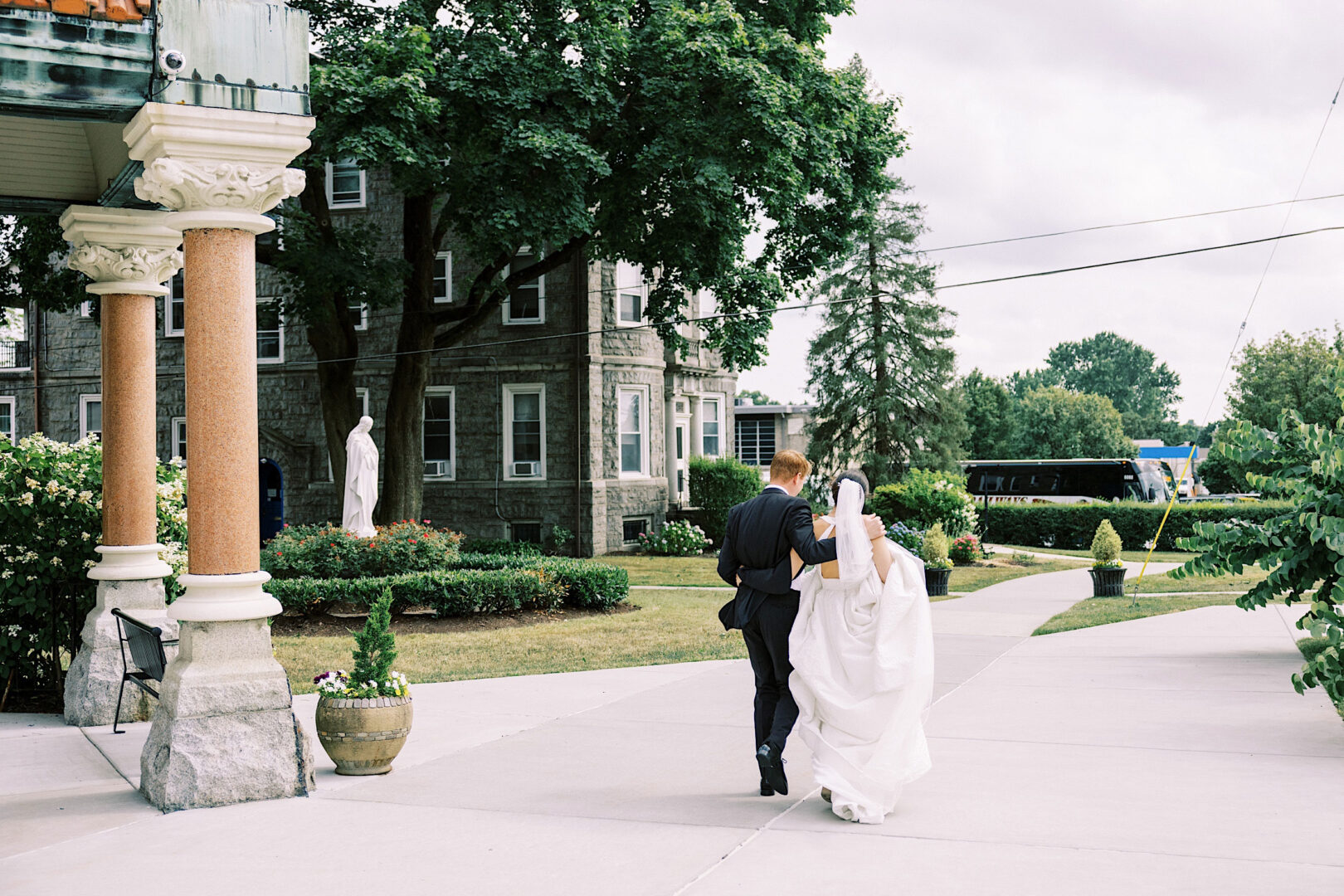 A couple dressed in wedding attire walks arm-in-arm on a path outside a stone building at Overbrook Golf Club in Villanova, surrounded by greenery and ornamental columns.